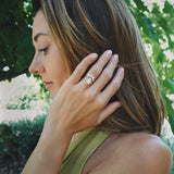 A woman in profile outdoors wearing a large halo-style ring with a central clear gemstone and surrounding metallic setting on her finger, with green foliage in the background.