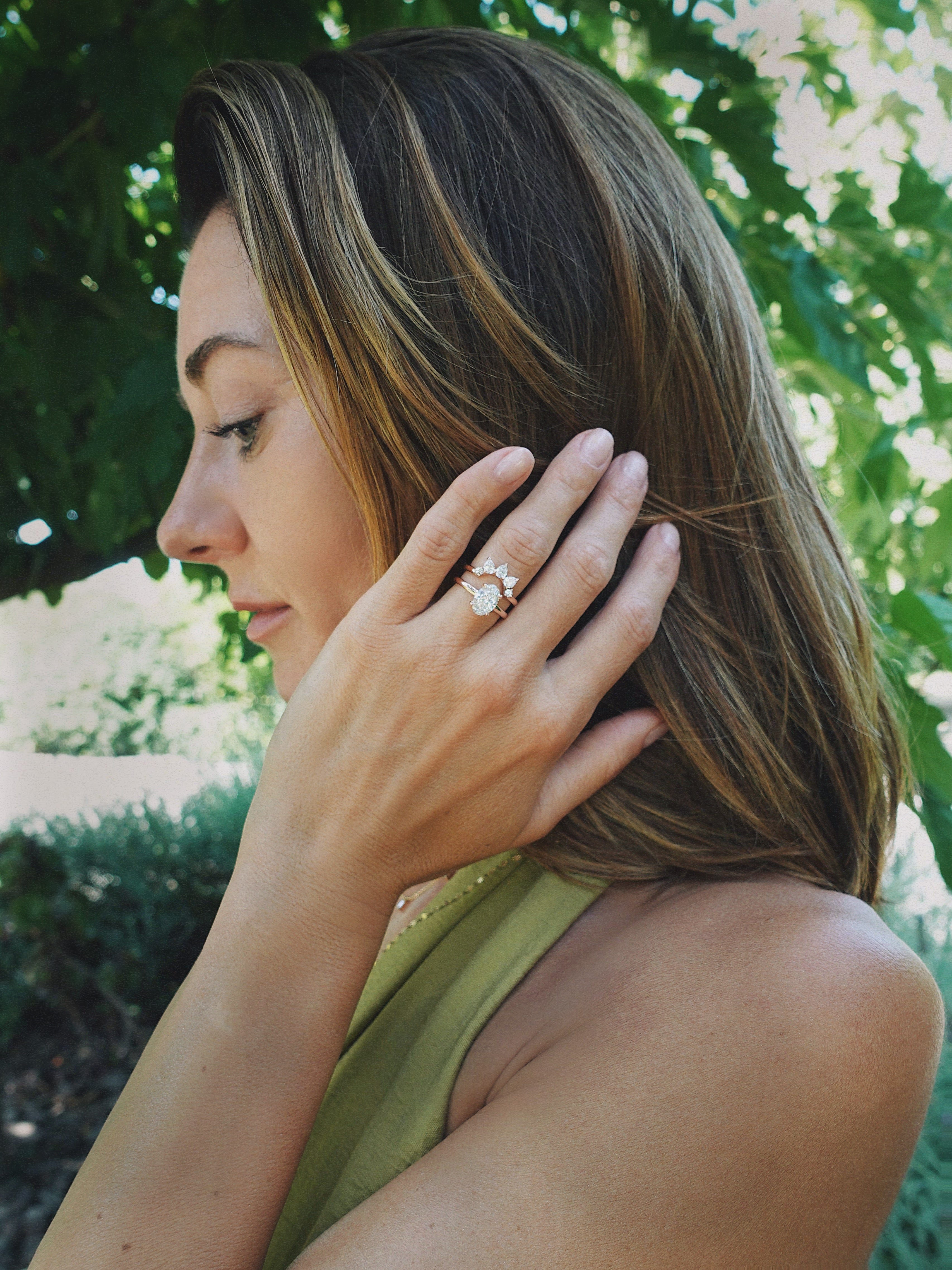 A woman in profile outdoors wearing a large halo-style ring with a central clear gemstone and surrounding metallic setting on her finger, with green foliage in the background.