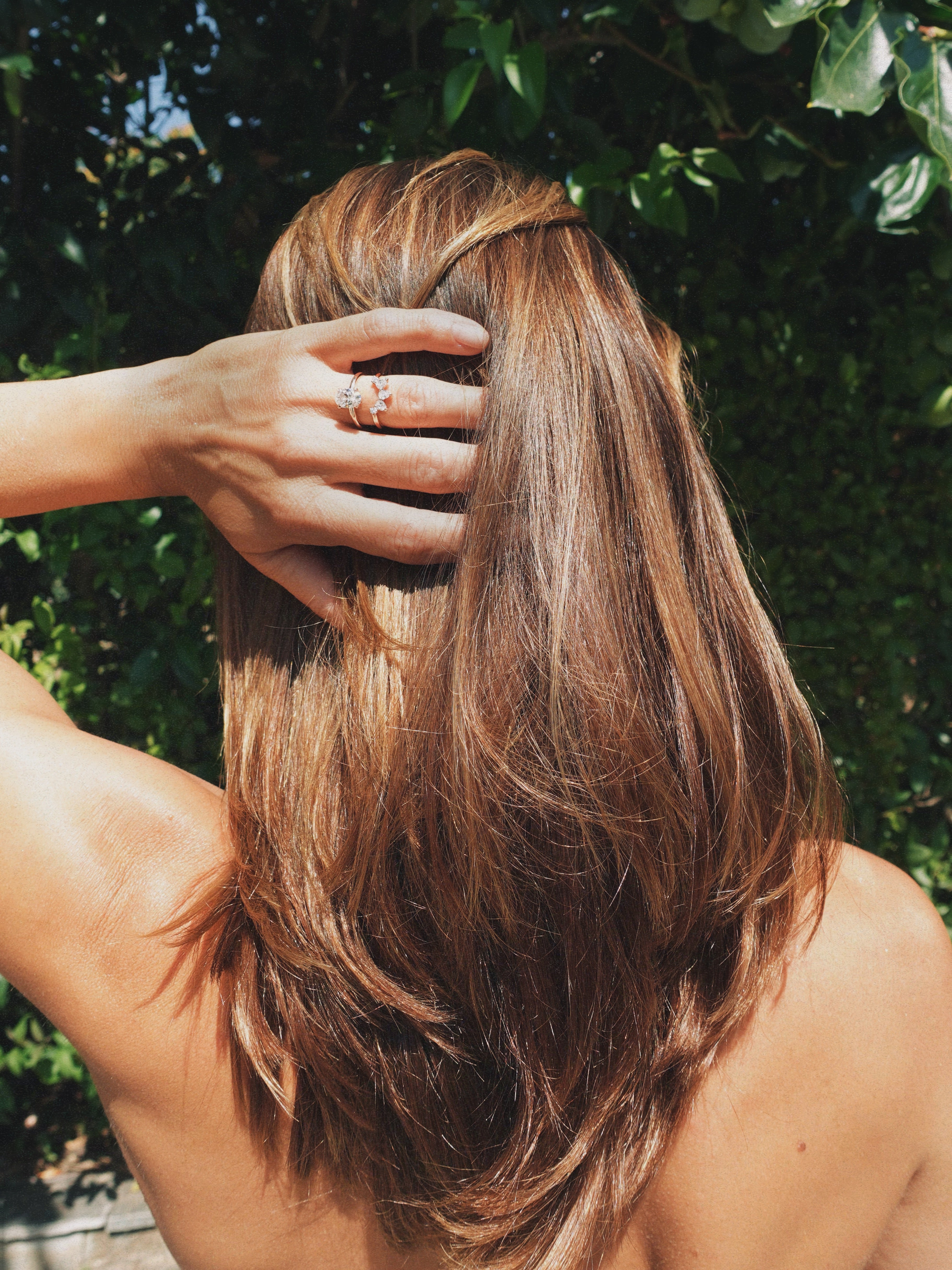 A woman outdoors with her back to the camera, showing shoulder-length brown hair with lighter warm highlights. Her hand is raised near her head wearing a beautiful engagement ring and matching band, and green foliage is visible in the background.