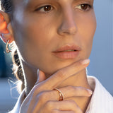 Close-up of a woman with her hand near her chin, wearing Papadopoulos gold diamond crossover hoops styled with a white shirt