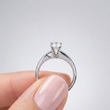 Diamond ring held by a hand with pink nail polish on a light gray background