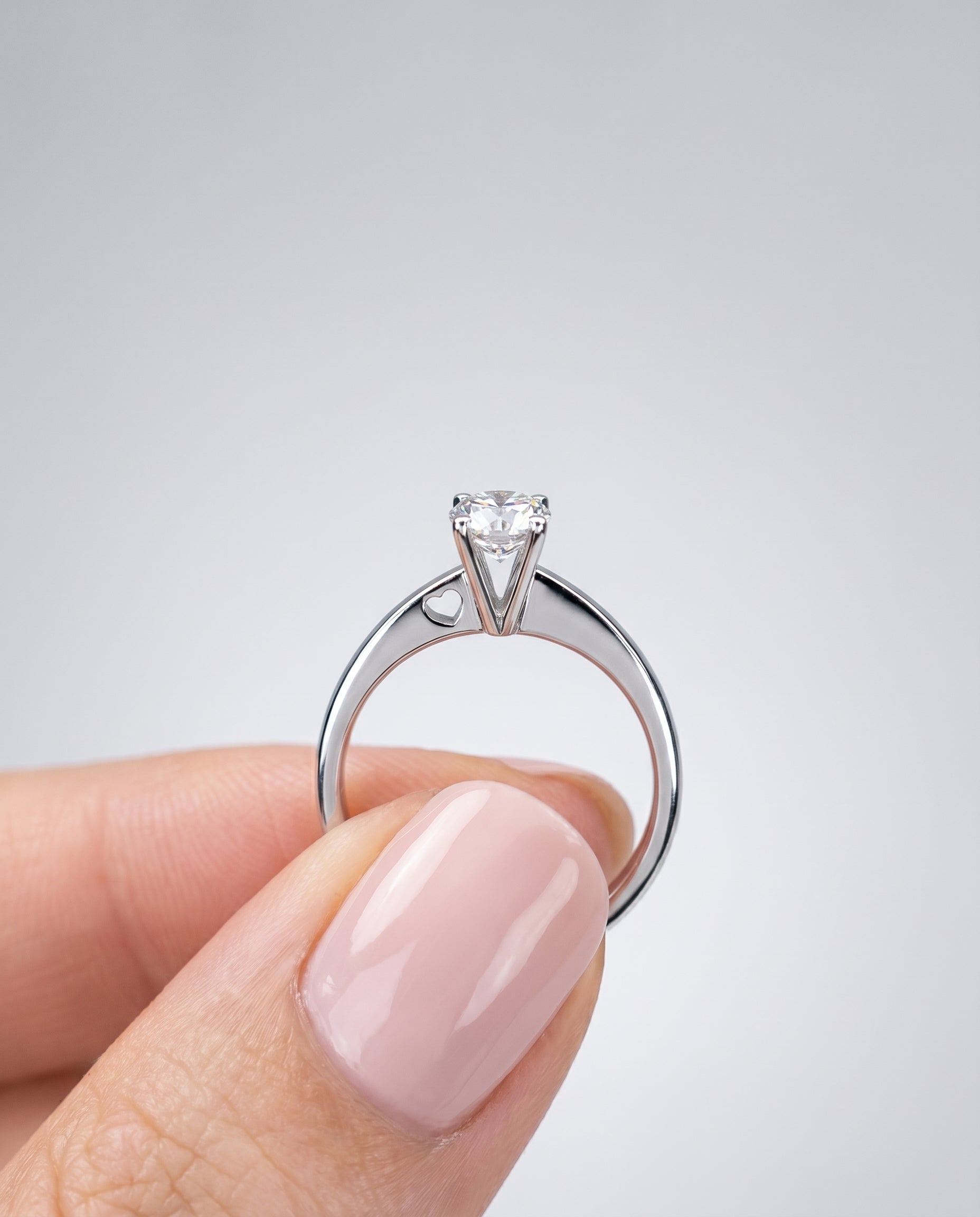 Diamond ring held by a hand with pink nail polish on a light gray background