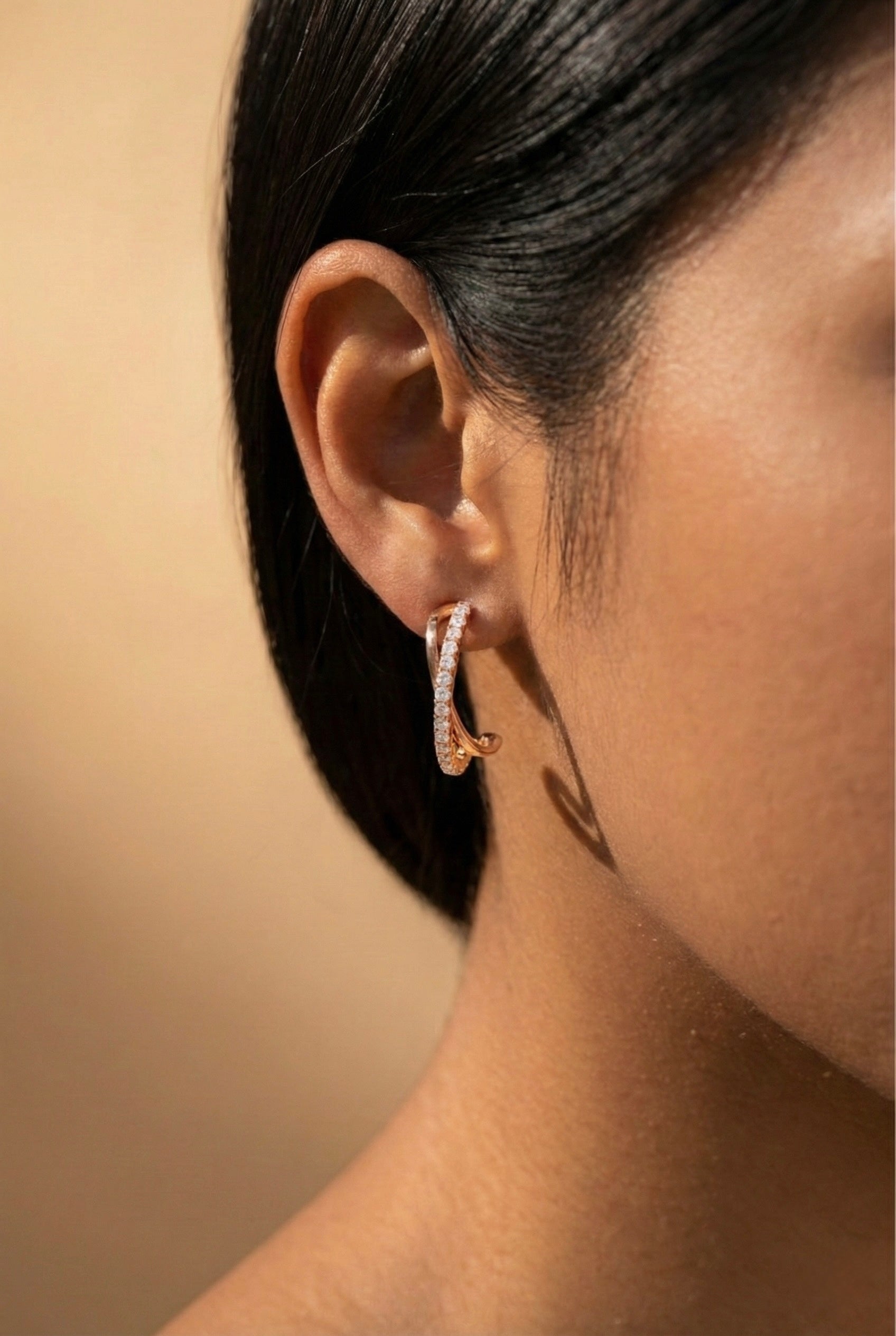 Close-up of an ear wearing a rose gold crossover hoop earring with a neutral background