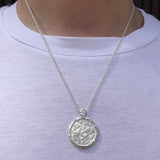 A silver pendant necklace with a textured circular medallion featuring Alexander the Great, worn on a person's neck over a white t-shirt.