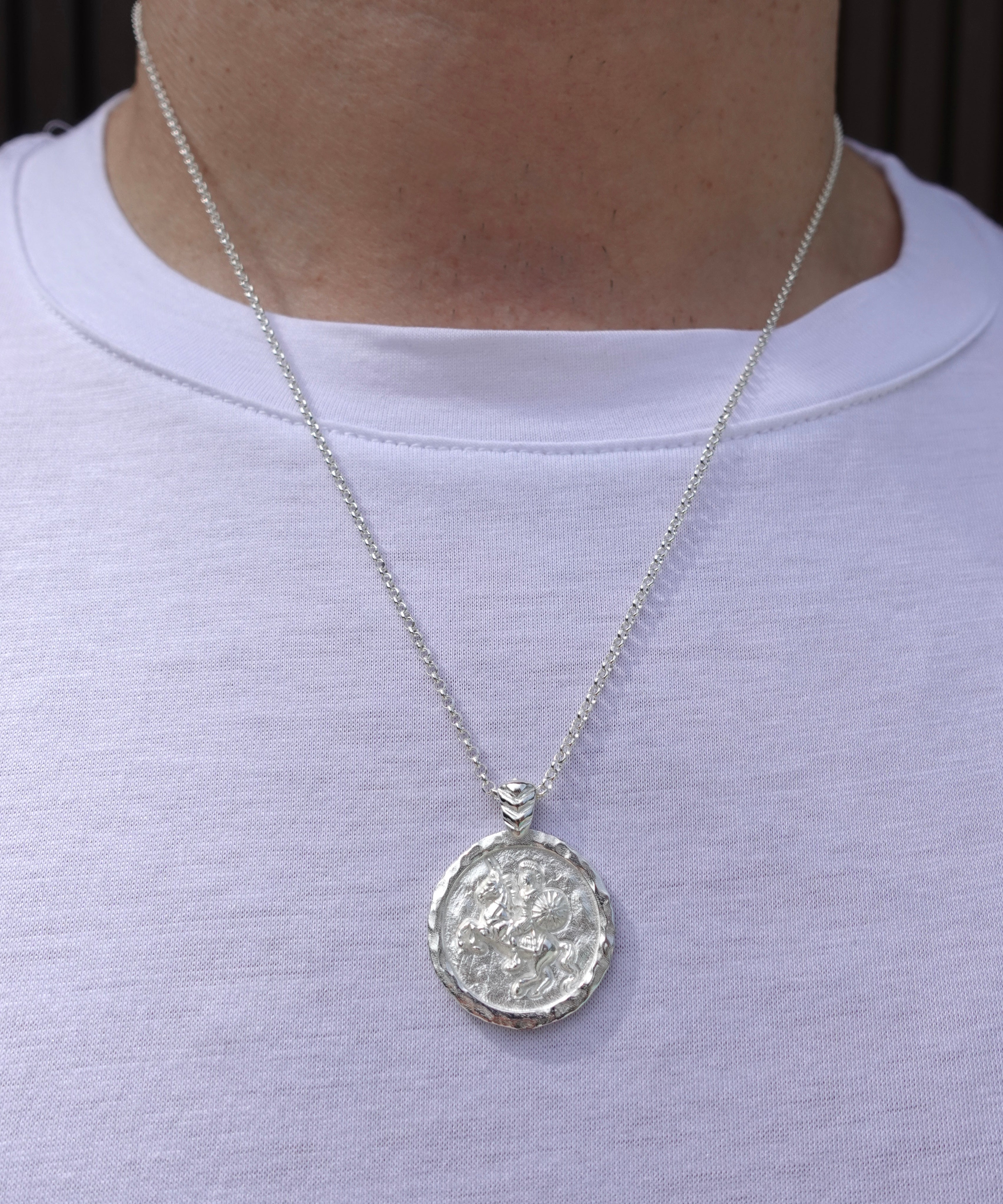 A silver pendant necklace with a textured circular medallion featuring Alexander the Great, worn on a person's neck over a white t-shirt.
