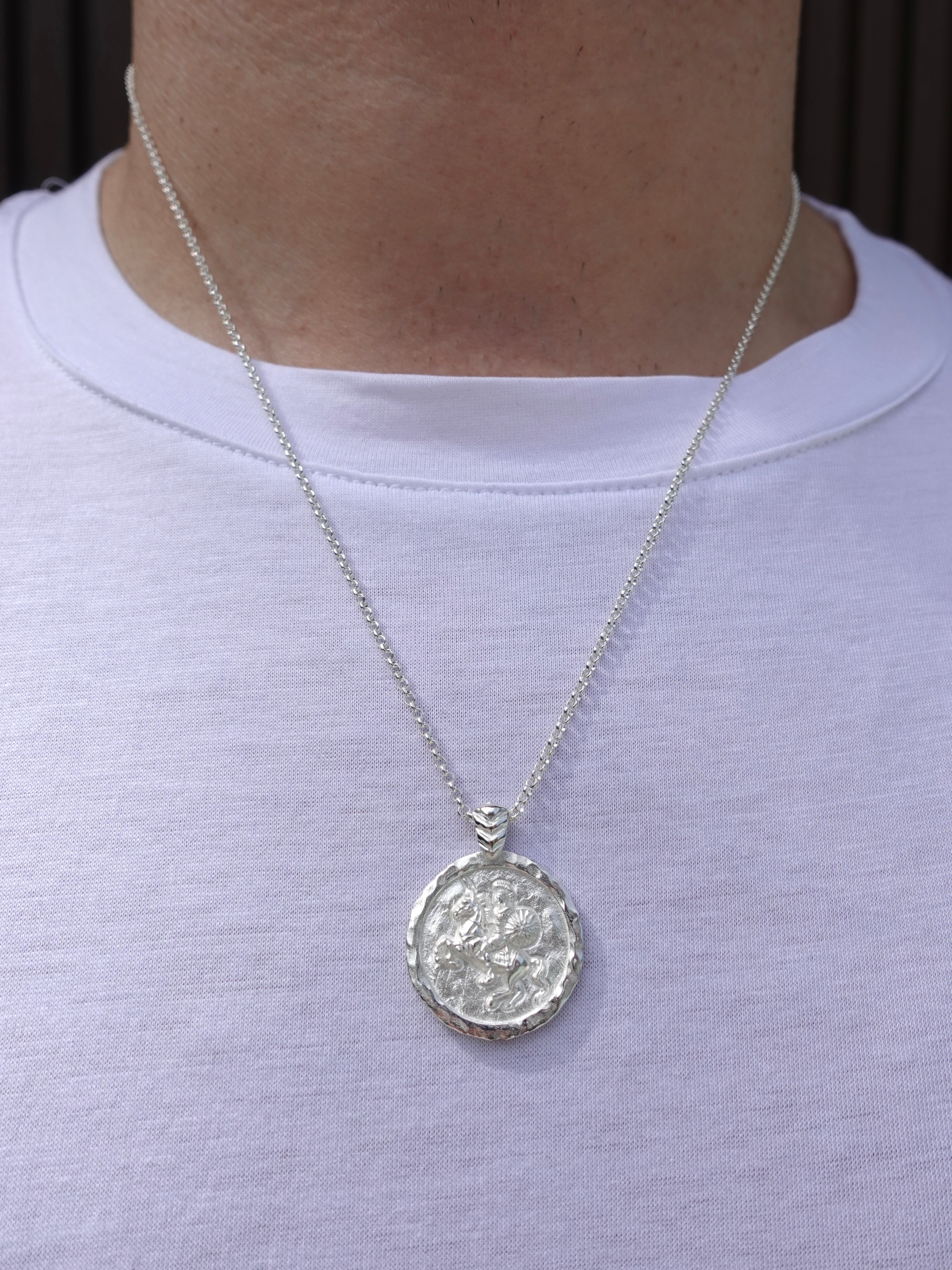 A silver pendant necklace with a textured circular medallion featuring Alexander the Great, worn on a person's neck over a white t-shirt.