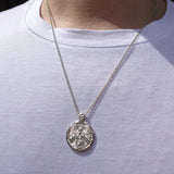 A silver pendant necklace with an embossed circular design, worn on a model's neck over a white t-shirt.