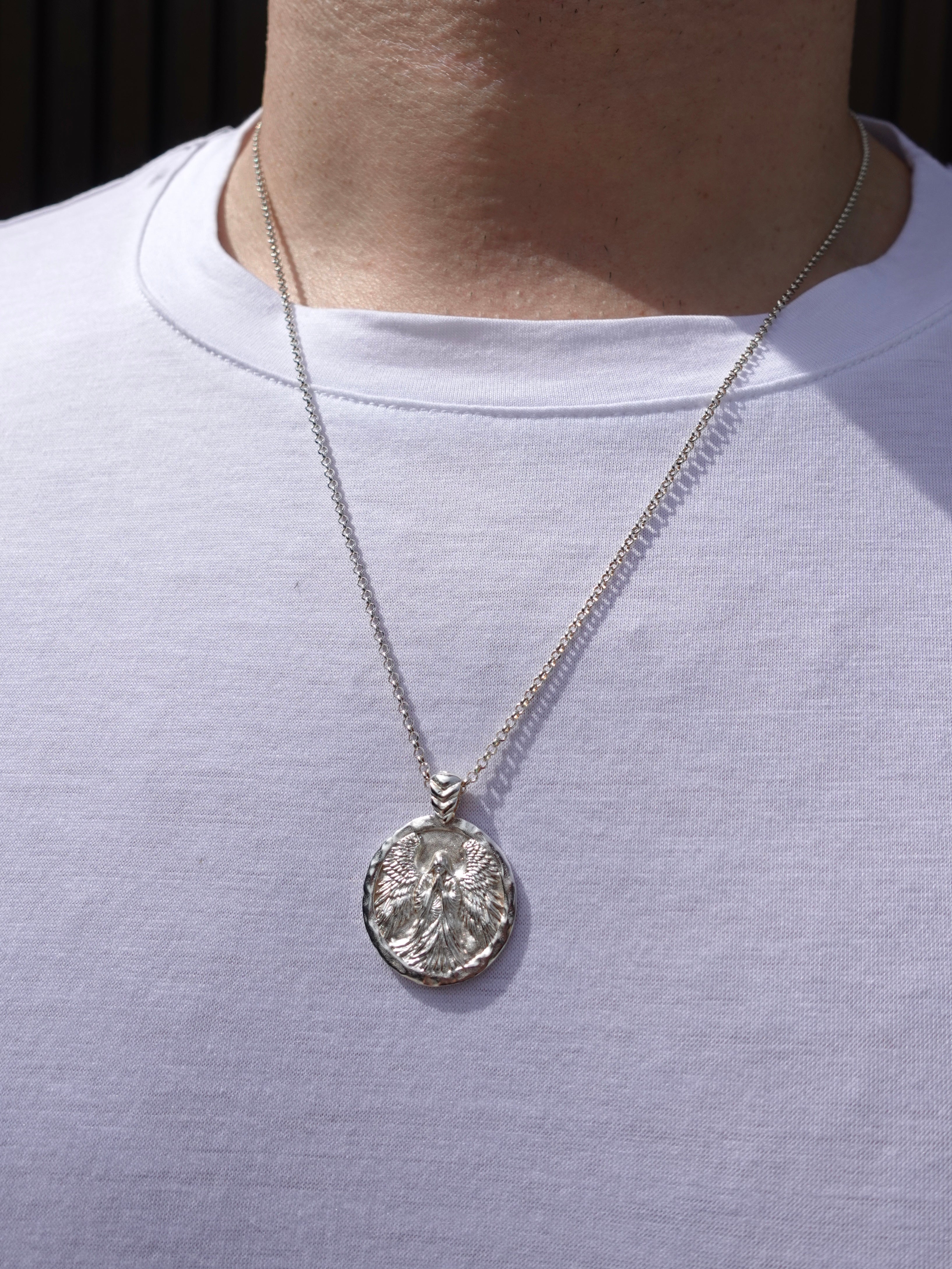 A silver pendant necklace with an embossed circular design, worn on a model's neck over a white t-shirt.