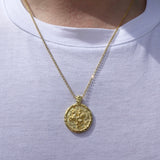 A gold coin pendant necklace worn by a person in a white t-shirt. The pendant features an embossed design resembling an ancient Roman or Greek coin featuring Alexander the Great.