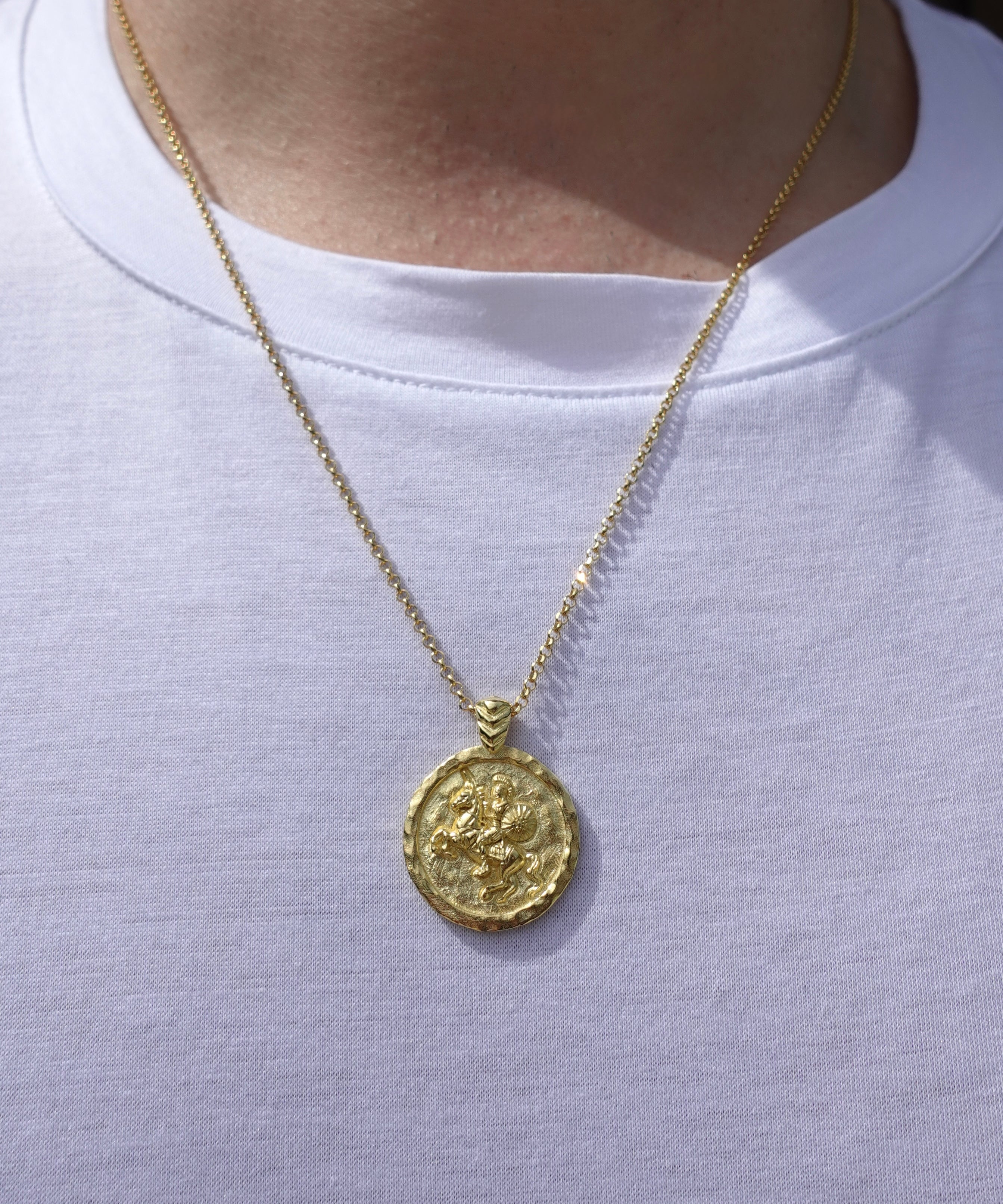 A gold coin pendant necklace worn by a person in a white t-shirt. The pendant features an embossed design resembling an ancient Roman or Greek coin featuring Alexander the Great.