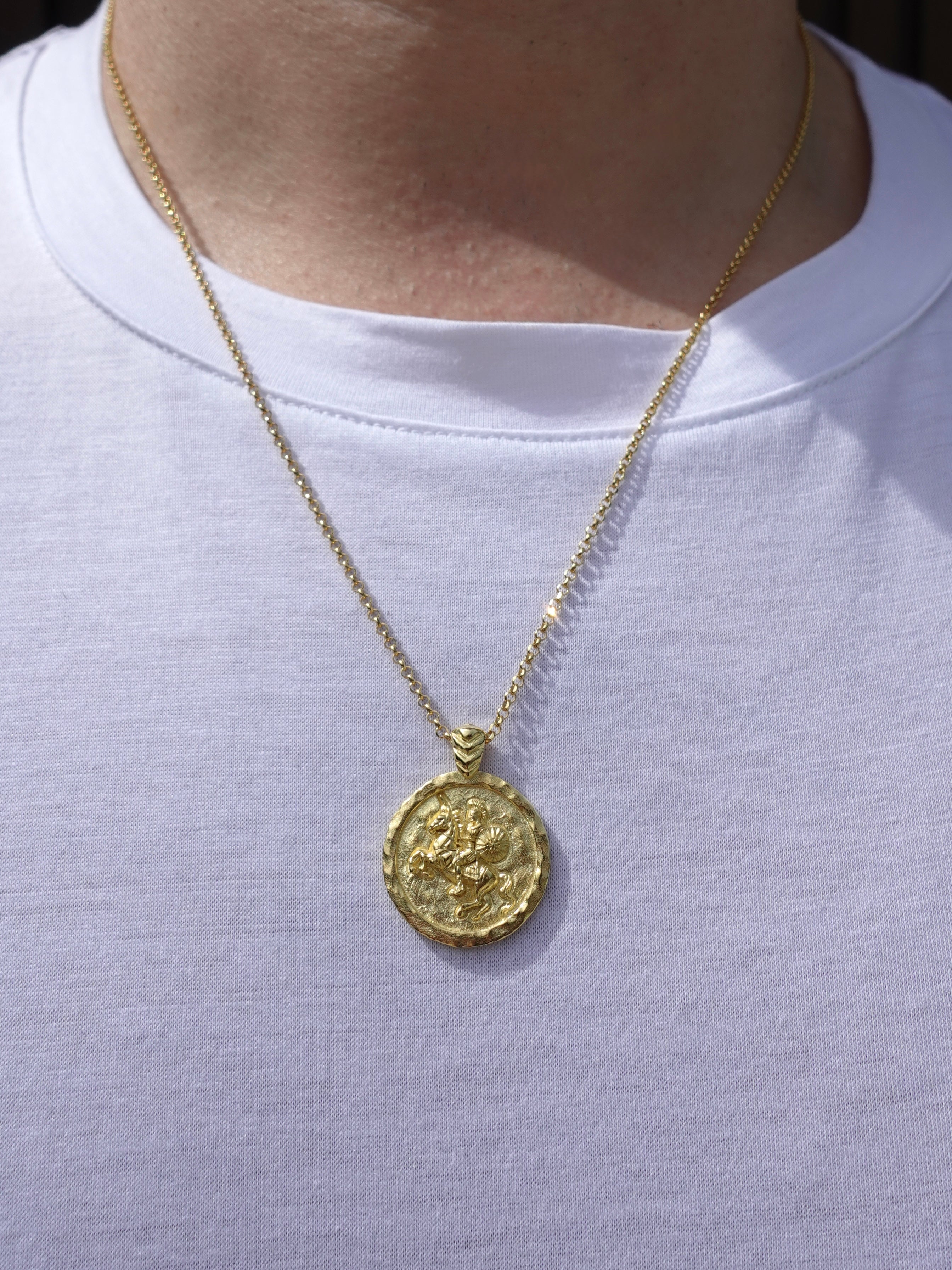 A gold coin pendant necklace worn by a person in a white t-shirt. The pendant features an embossed design resembling an ancient Roman or Greek coin featuring Alexander the Great.