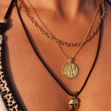 A close-up of a person's neck and chest wearing three layered necklaces: a gold chain with a round coin pendant, a gold chain with gold coin design with angel, and a black cord necklace with a ancient greek helmet. 