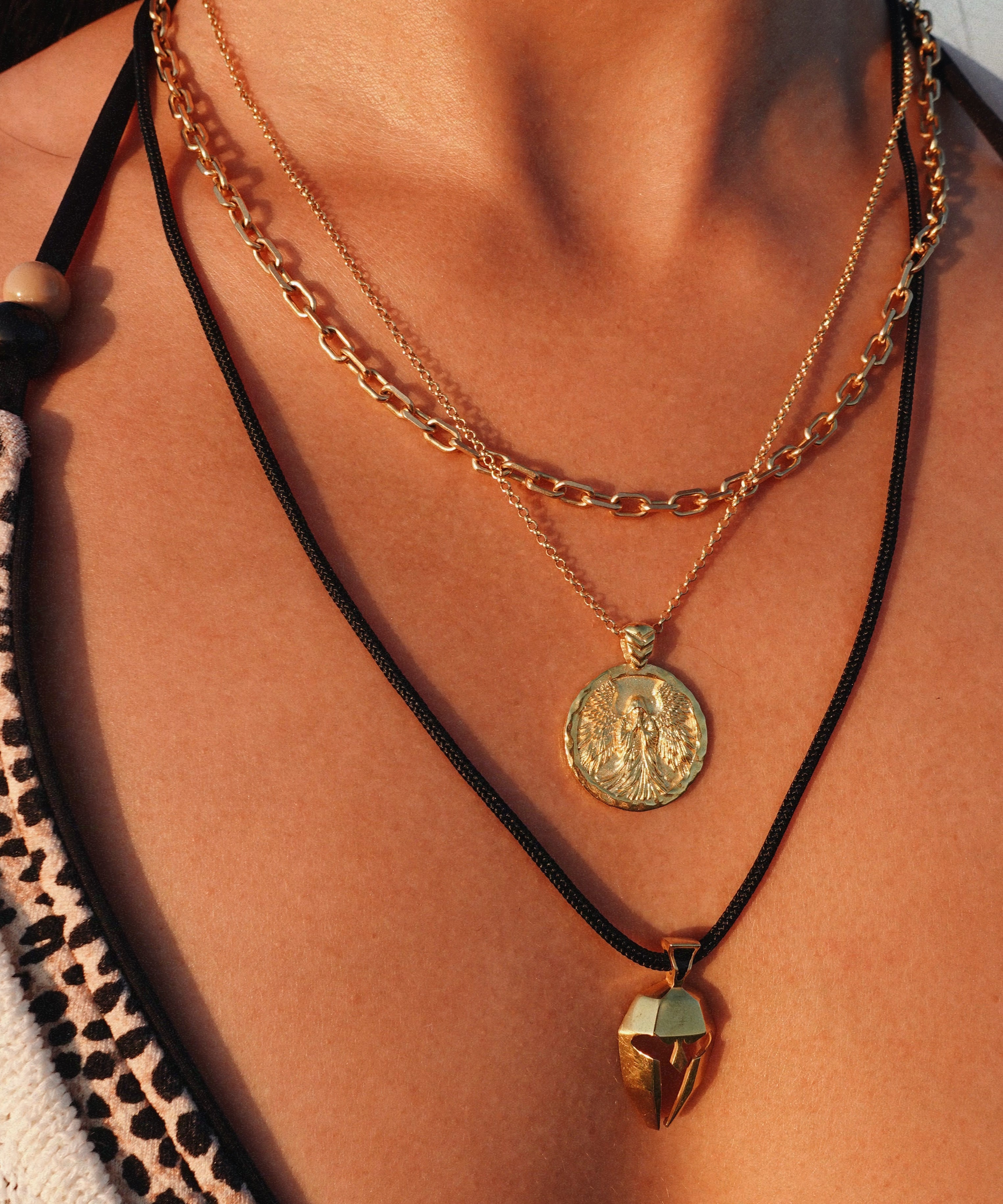 A close-up of a person's neck and chest wearing three layered necklaces: a gold chain with a round coin pendant, a gold chain with gold coin design with angel, and a black cord necklace with a ancient greek helmet. 