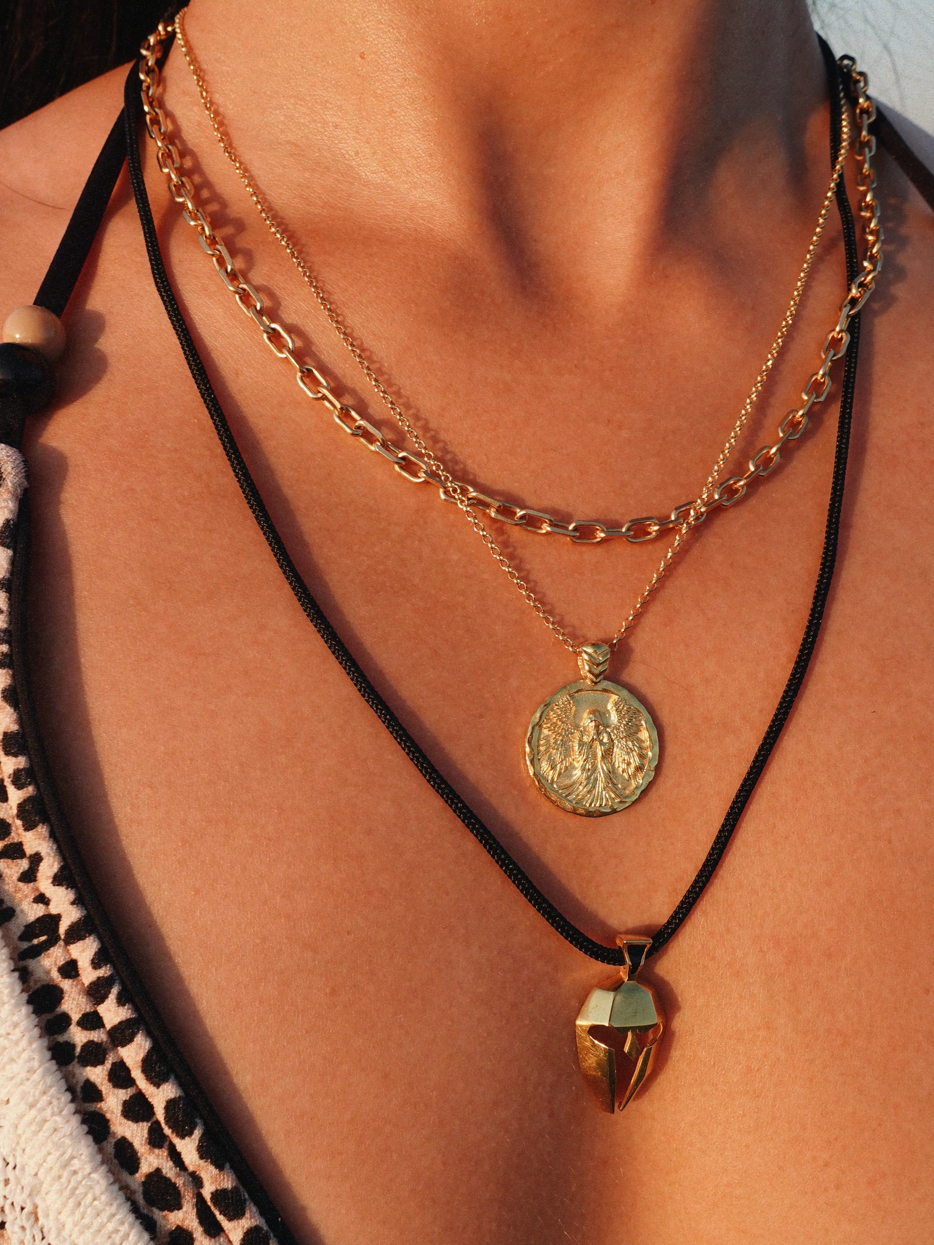 A close-up of a person's neck and chest wearing three layered necklaces: a gold chain with a round coin pendant, a gold chain with gold coin design with angel, and a black cord necklace with a ancient greek helmet. 