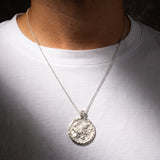 A silver pendant necklace worn by a person in a white t-shirt, featuring a circular textured pendant with an embossed griffin design.