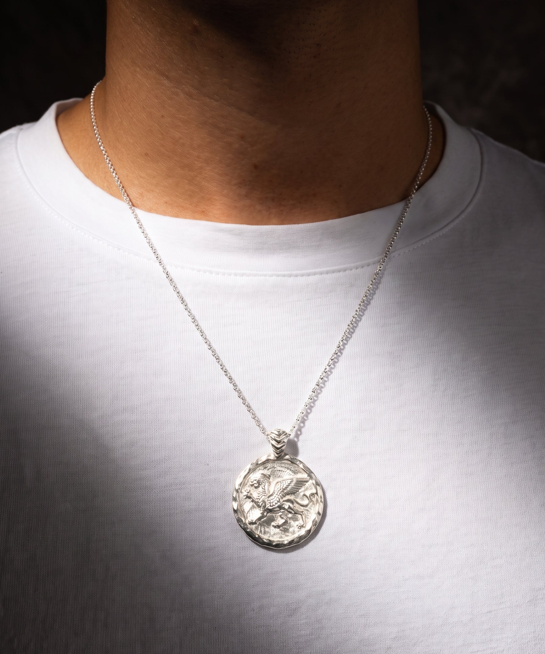 A silver pendant necklace worn by a person in a white t-shirt, featuring a circular textured pendant with an embossed griffin design.