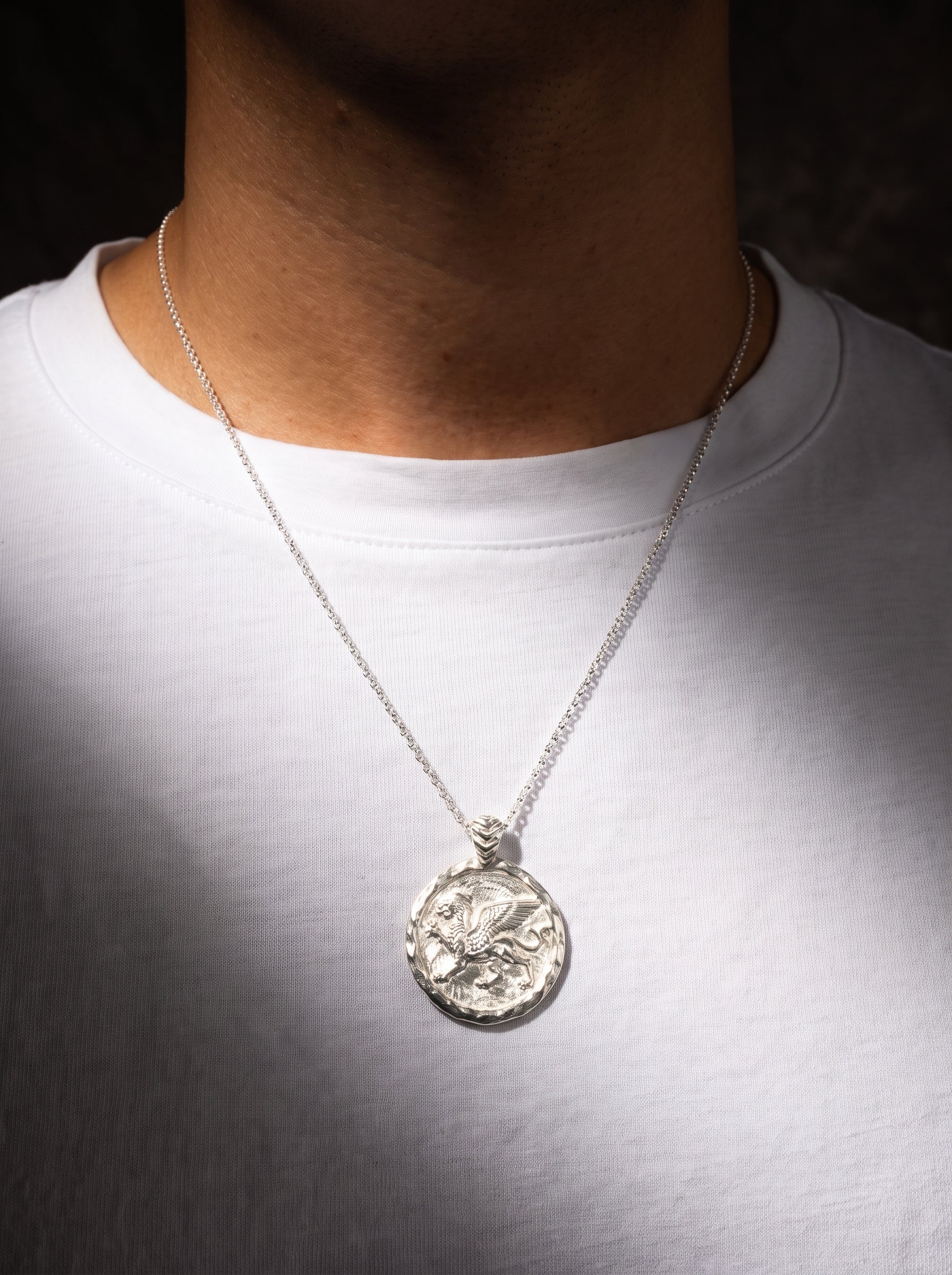 A silver pendant necklace worn by a person in a white t-shirt, featuring a circular textured pendant with an embossed griffin design.