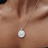 A silver coin pendant necklace worn on a model's neck, featuring a textured circular pendant on a delicate silver chain.