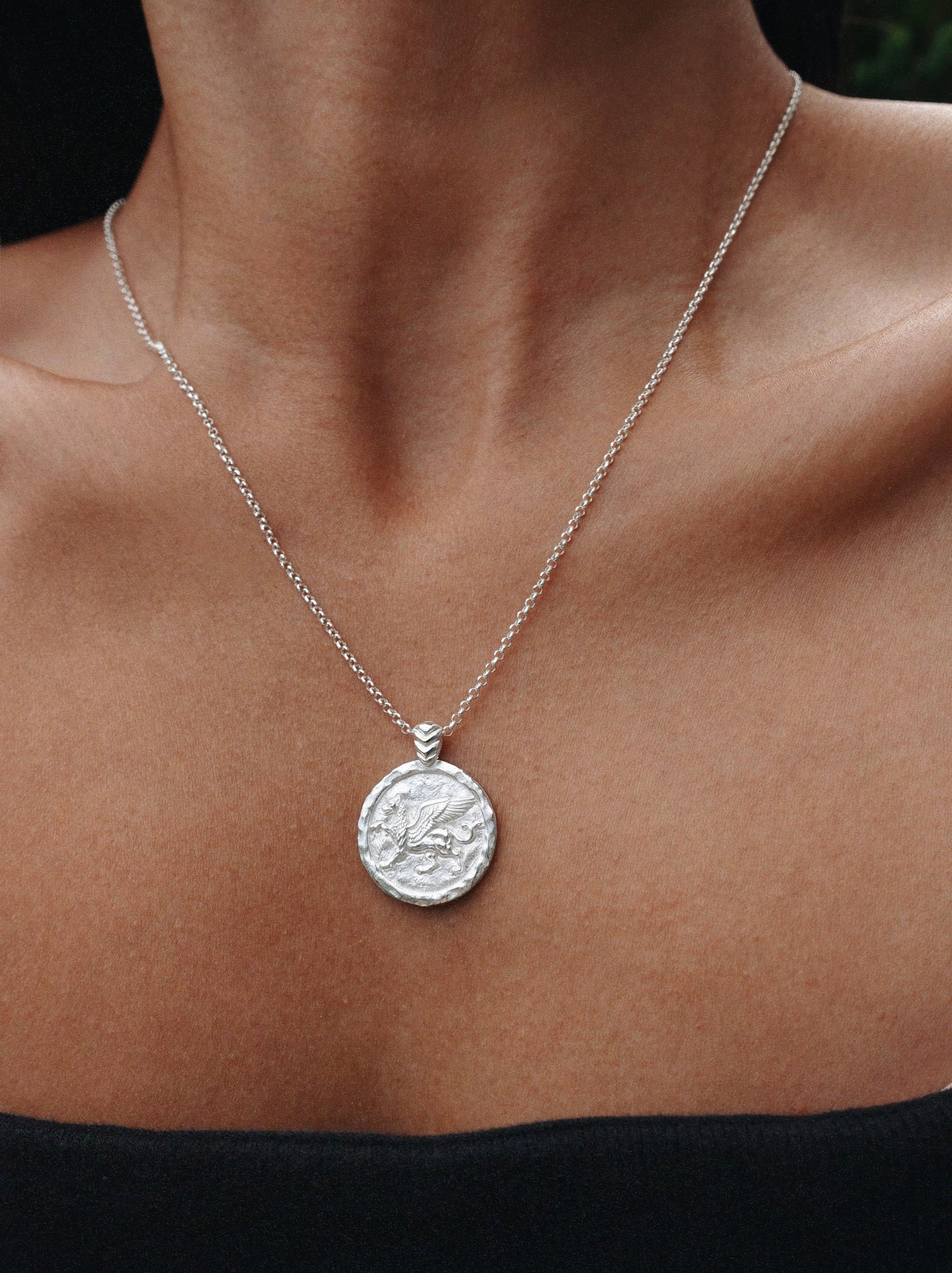 A silver coin pendant necklace worn on a model's neck, featuring a textured circular pendant on a delicate silver chain.