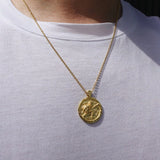 A gold coin pendant necklace with a warrior design worn on a person's neck over a white t-shirt.