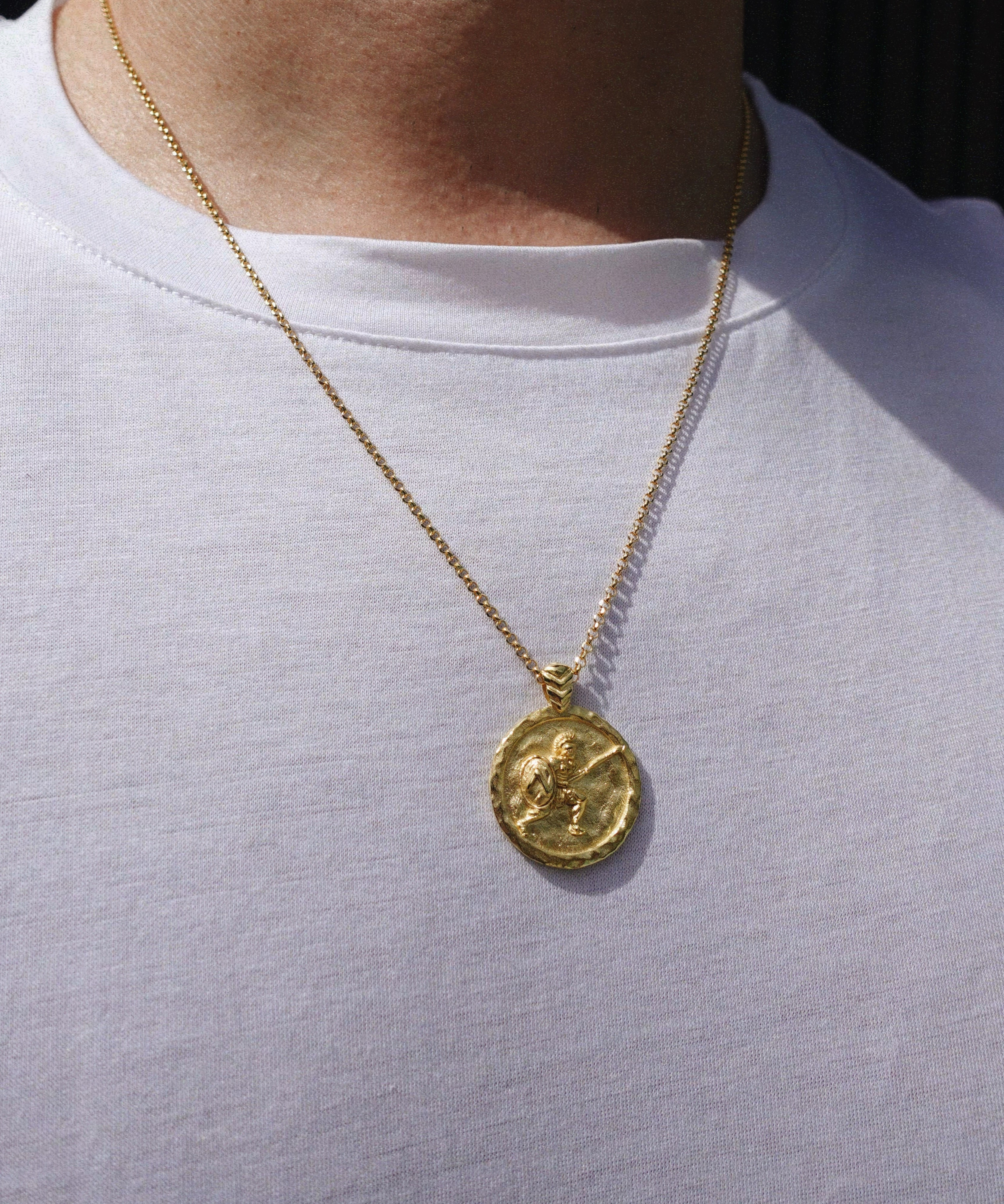 A gold coin pendant necklace with a warrior design worn on a person's neck over a white t-shirt.