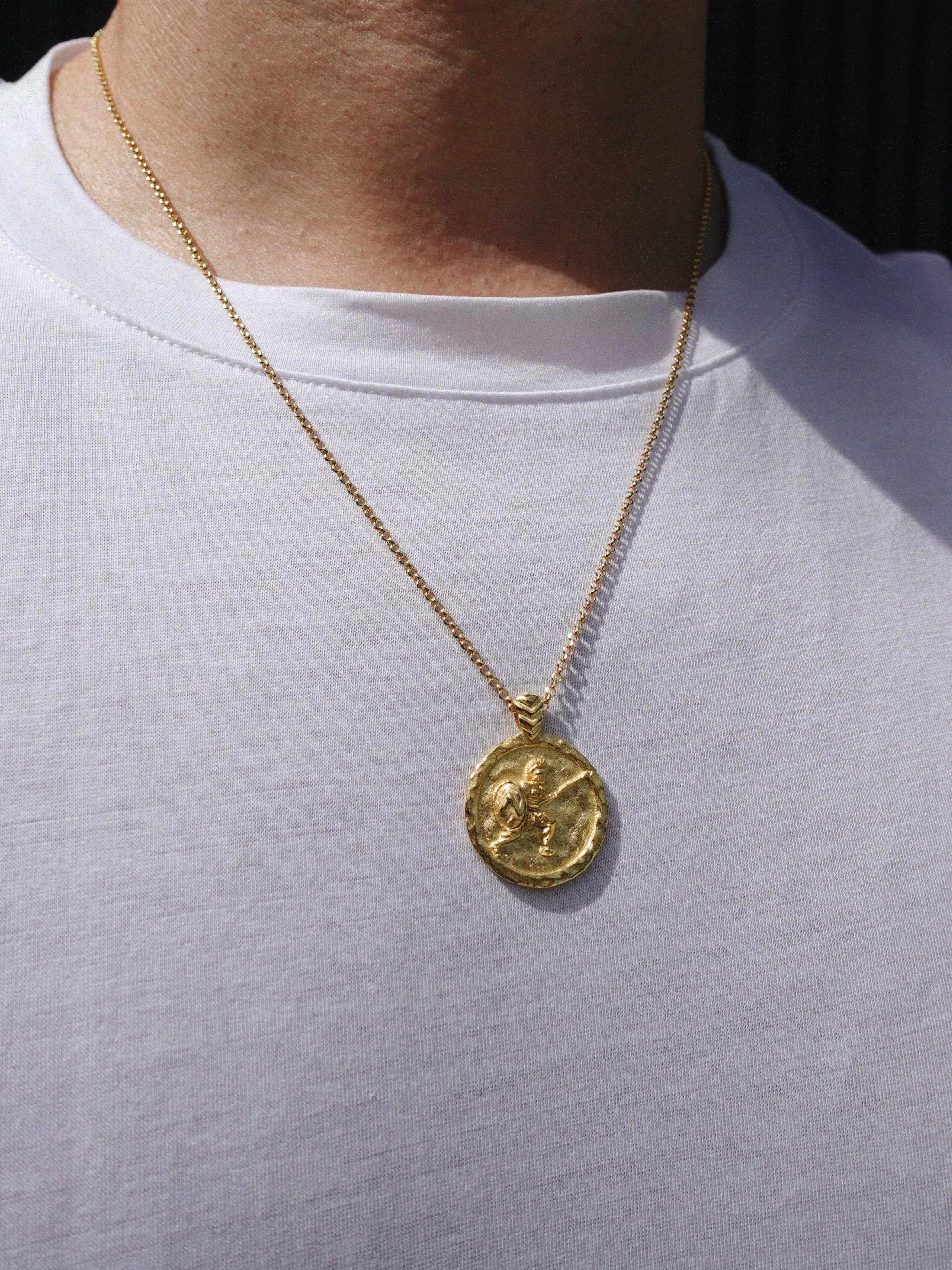 A gold coin pendant necklace with a warrior design worn on a person's neck over a white t-shirt.