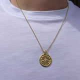 A gold pendant necklace with a textured circular medallion and a small clear stone, worn by a person in a white t-shirt.