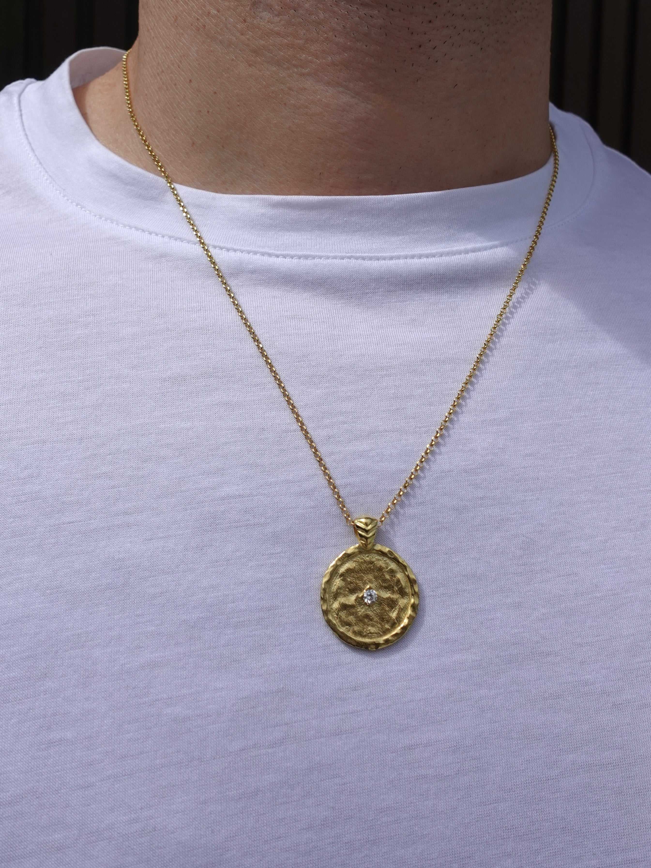 A gold pendant necklace with a textured circular medallion and a small clear stone, worn by a person in a white t-shirt.