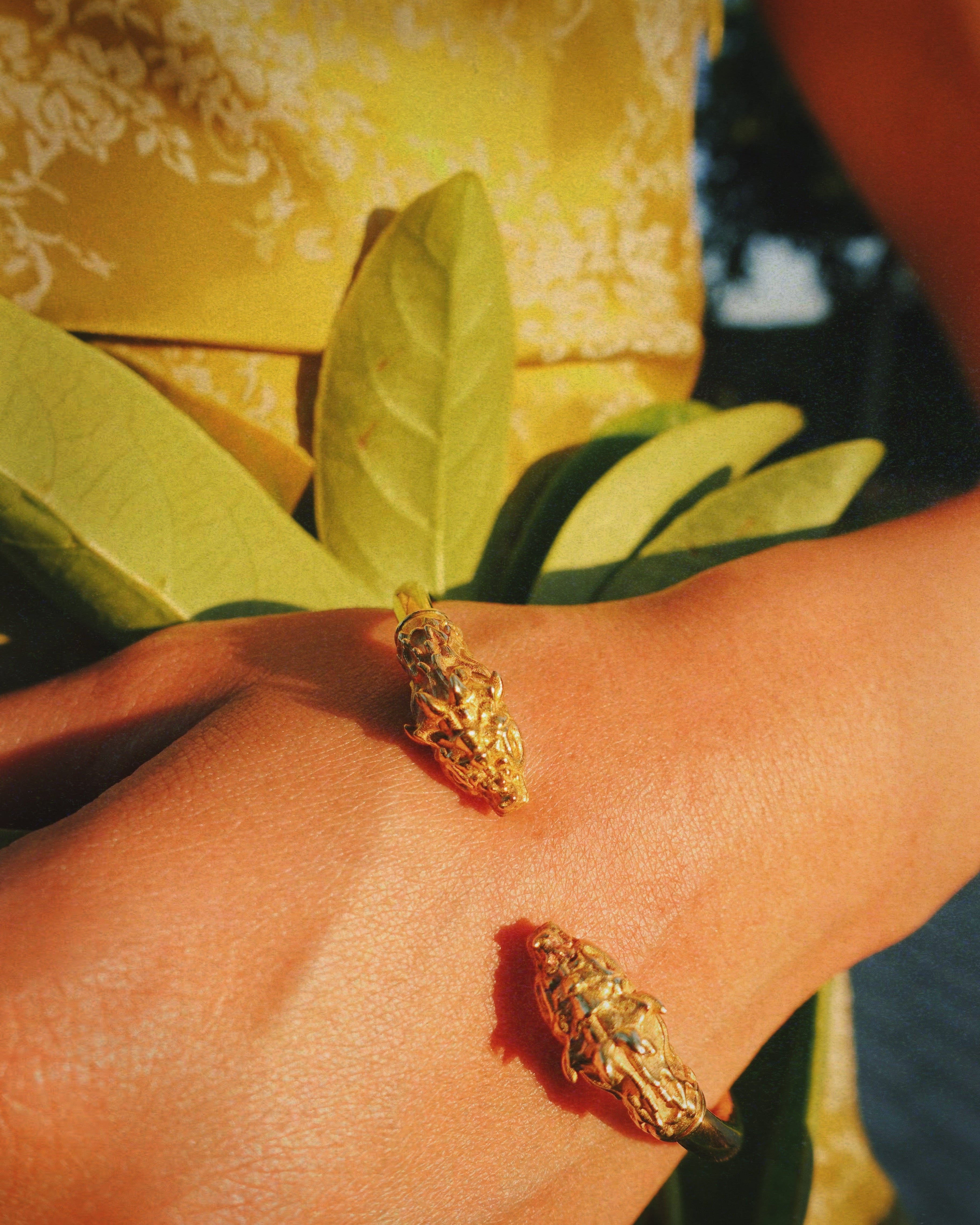 A gold-toned dragon head bracelet worn on a person's wrist, with a yellow patterned garment and green leaves in the background.