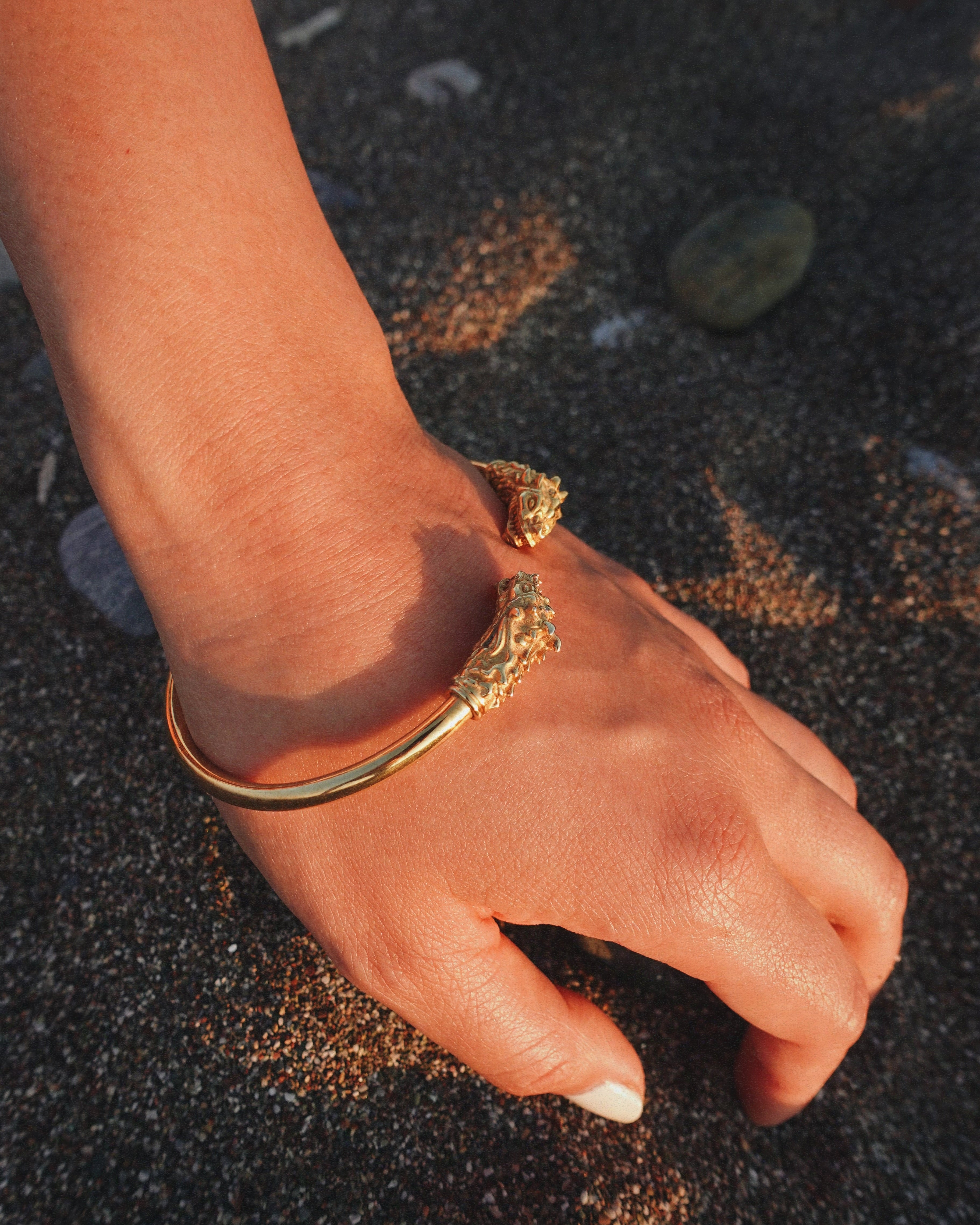 A gold-toned bangle bracelet with ornate dragon head details on the ends, worn on a person's wrist over dark sand.