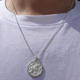 A silver pendant necklace with a textured, coin-like circular charm featuring a winged figure griffin, shown worn on a person's neck over a white t-shirt.