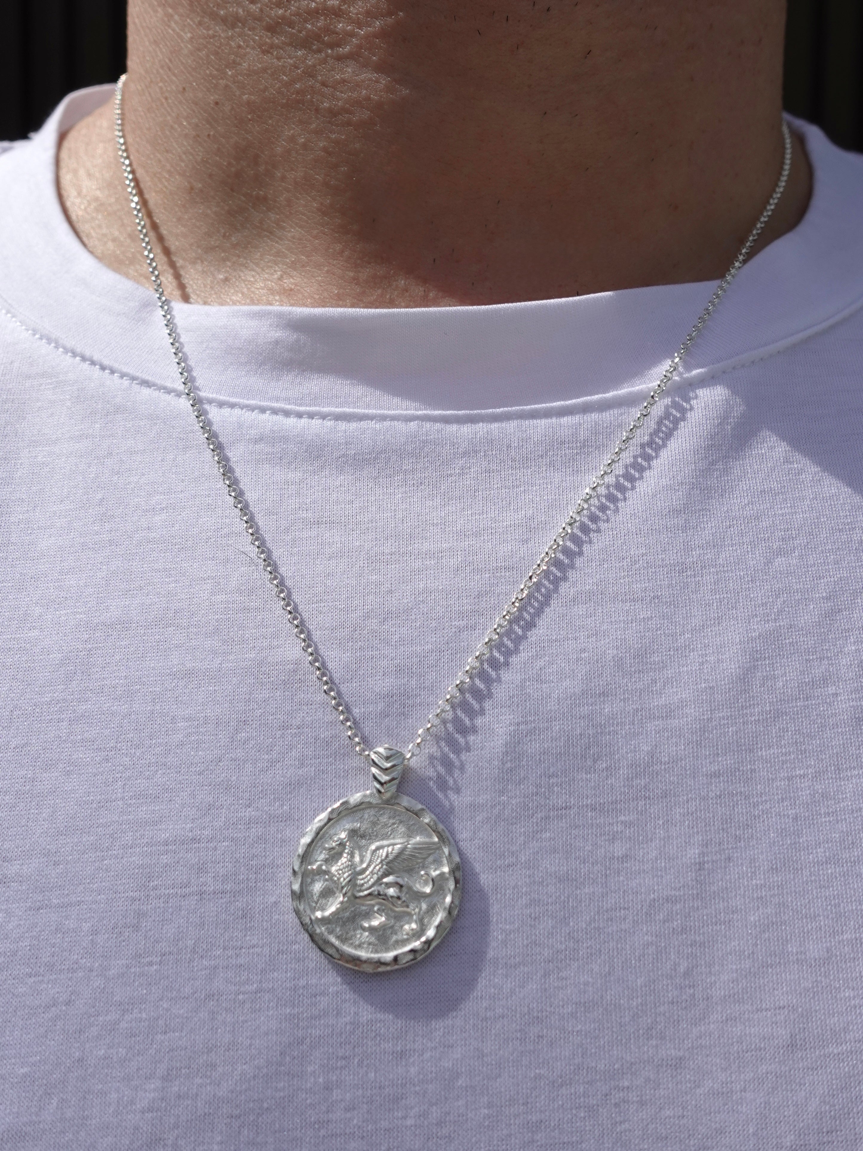 A silver pendant necklace with a textured, coin-like circular charm featuring a winged figure griffin, shown worn on a person's neck over a white t-shirt.