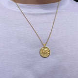 A gold coin pendant necklace worn by a person wearing a white t-shirt. The pendant features an embossed design resembling a classical coin with a small hanging loop at the top.