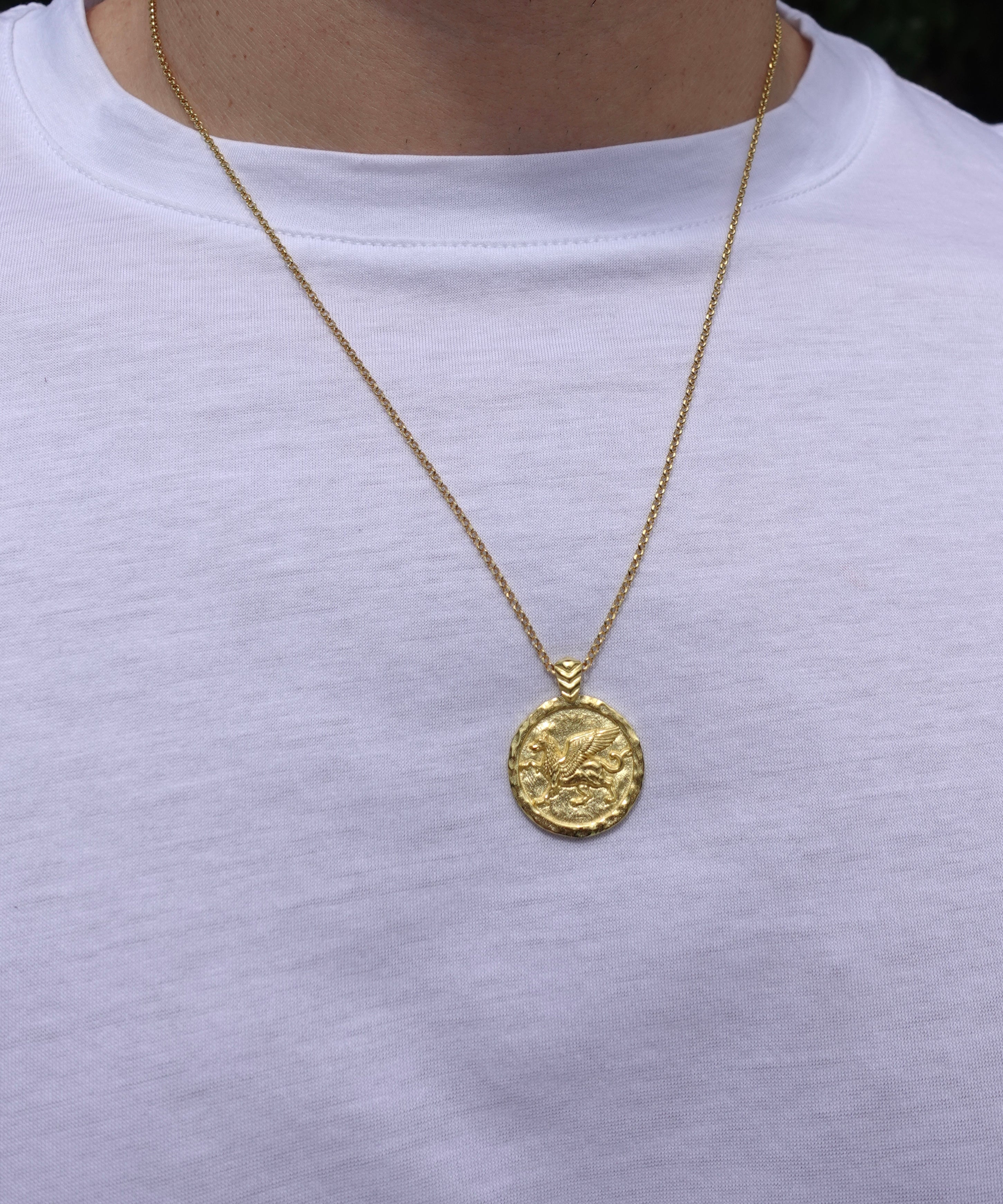 A gold coin pendant necklace worn by a person wearing a white t-shirt. The pendant features an embossed design resembling a classical coin with a small hanging loop at the top.