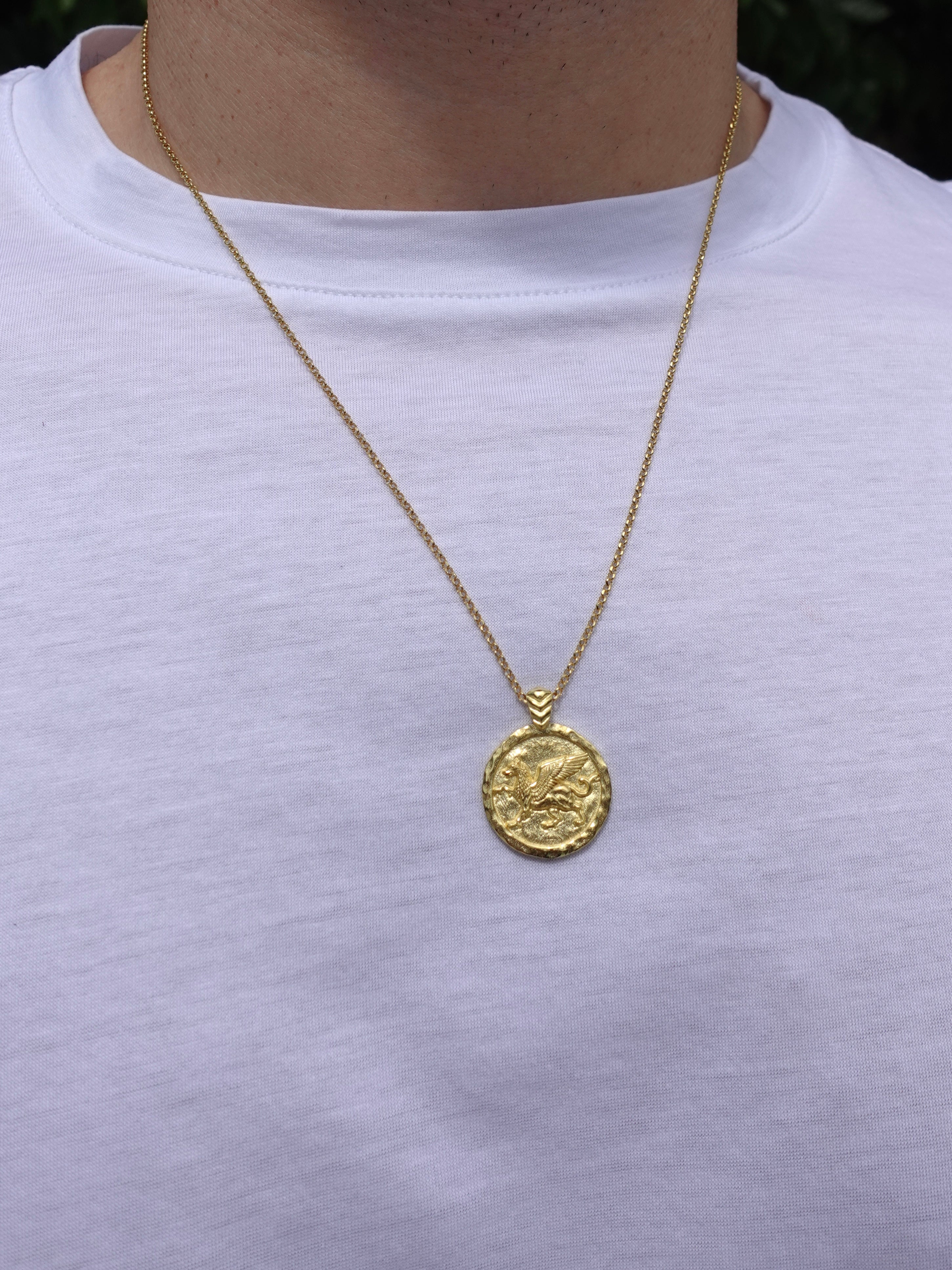 A gold coin pendant necklace worn by a person wearing a white t-shirt. The pendant features an embossed design resembling a classical coin with a small hanging loop at the top.