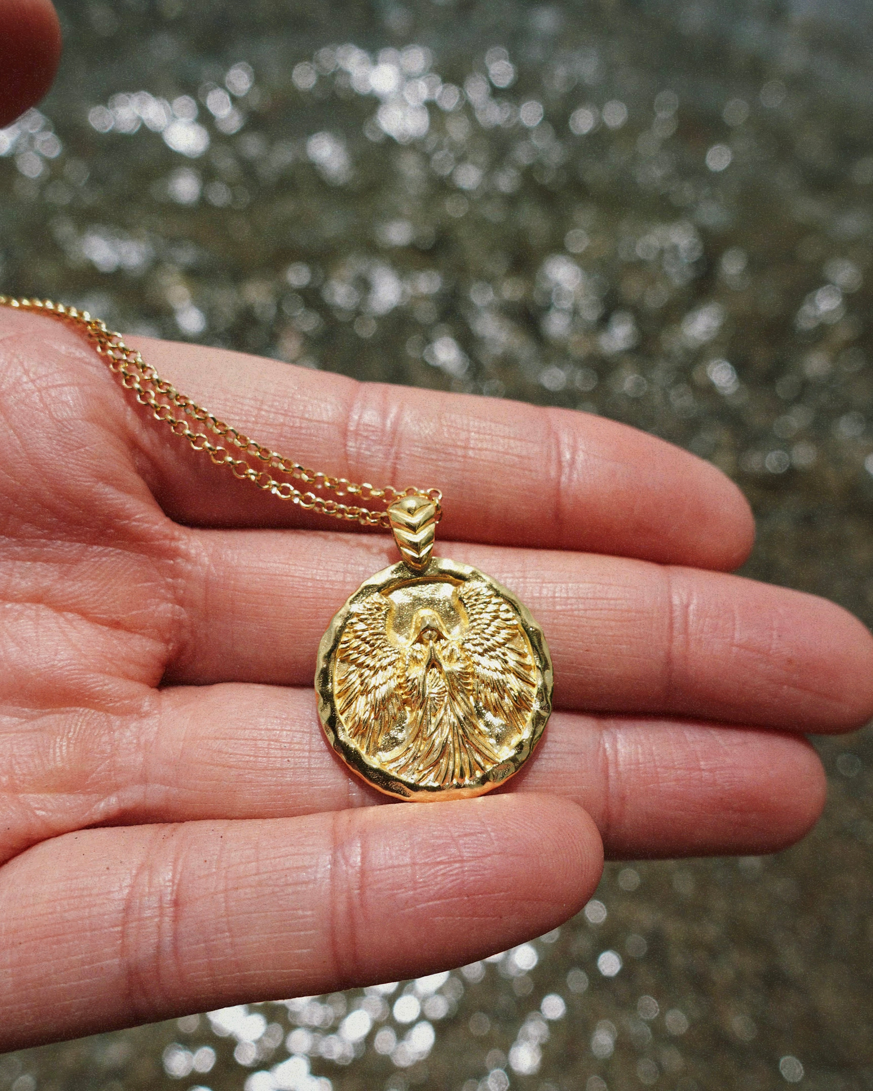 Gold necklace with a detailed pendant featuring a hooded angel held in a hand against a blurred water background