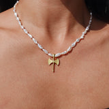 A close-up of a person's neck and upper chest wearing a necklace with irregular white pearl-like beads and a small gold axe pendant at the center.
