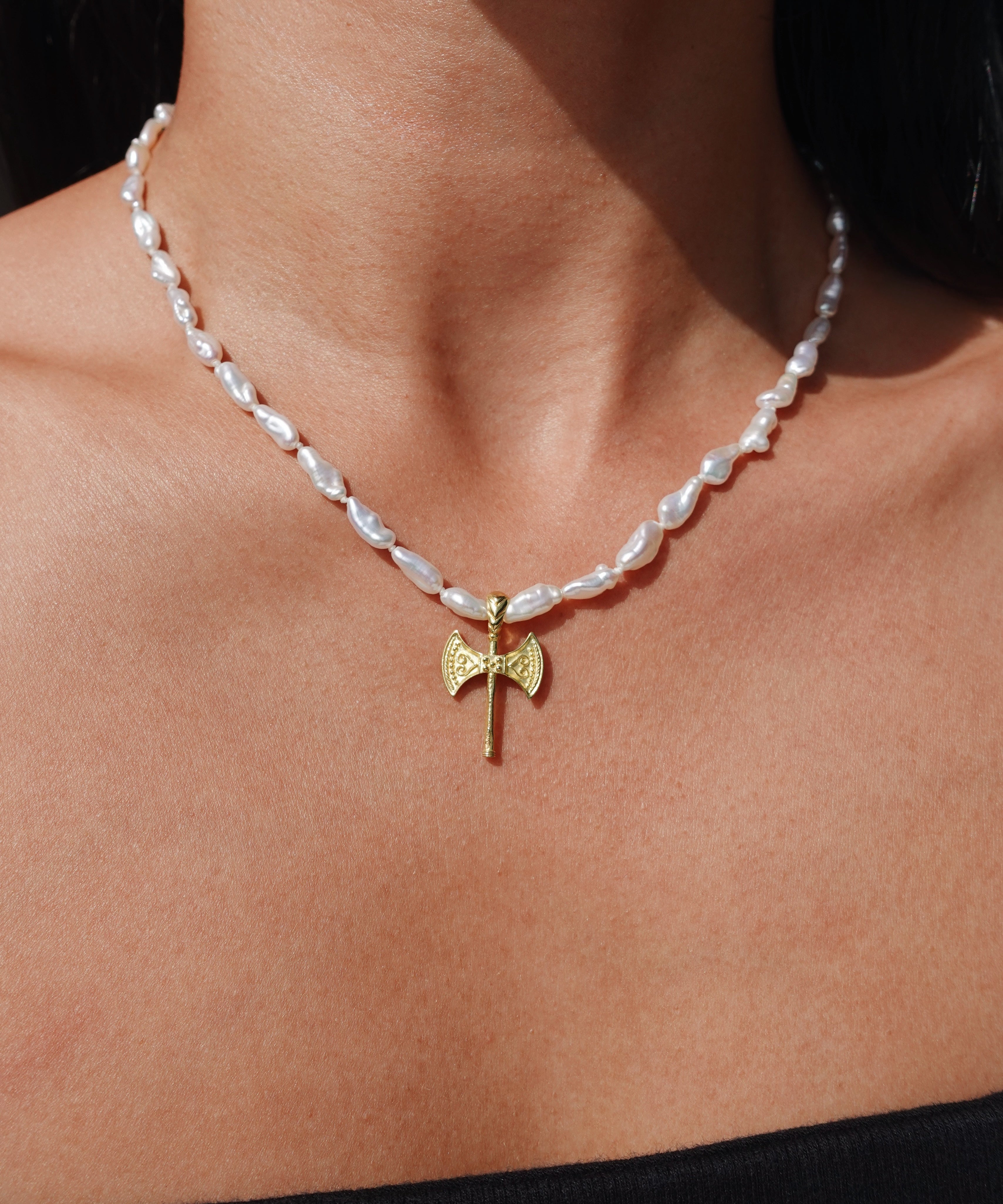 A close-up of a person's neck and upper chest wearing a necklace with irregular white pearl-like beads and a small gold axe pendant at the center.