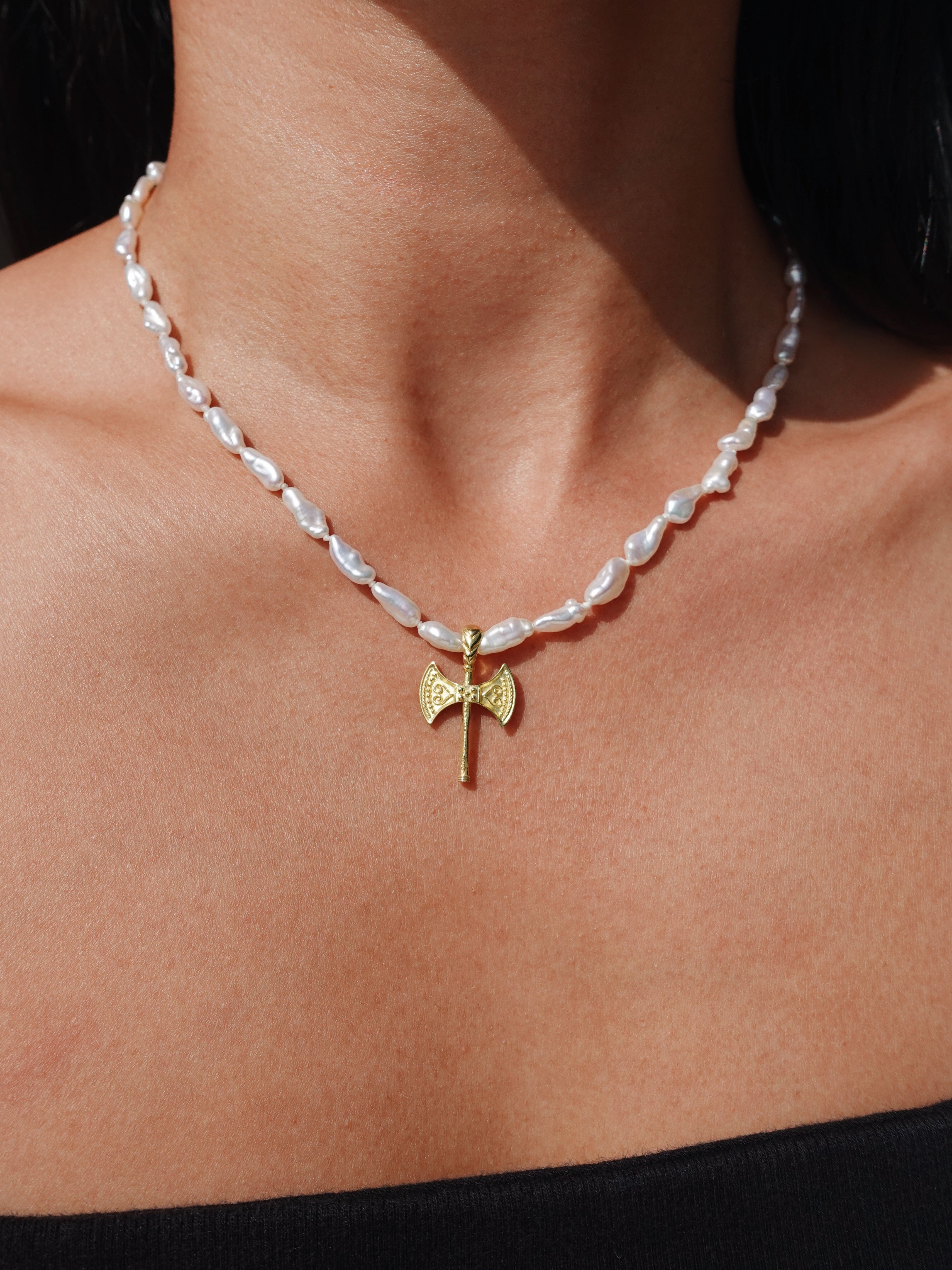 A close-up of a person's neck and upper chest wearing a necklace with irregular white pearl-like beads and a small gold axe pendant at the center.
