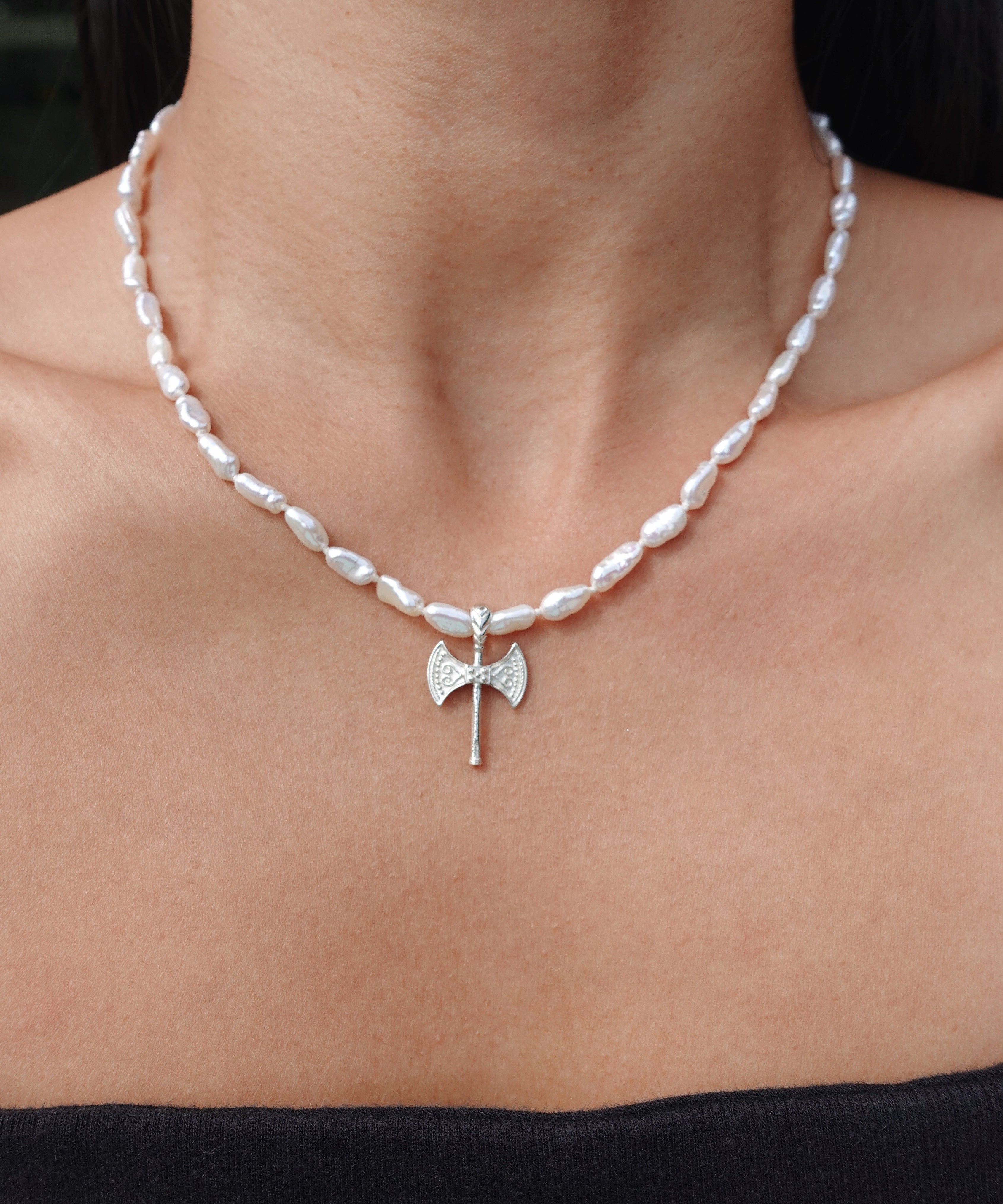 A close-up of a person's neck and upper chest wearing a silver necklace with irregular white pearl-like beads and a small axe pendant at the center.
