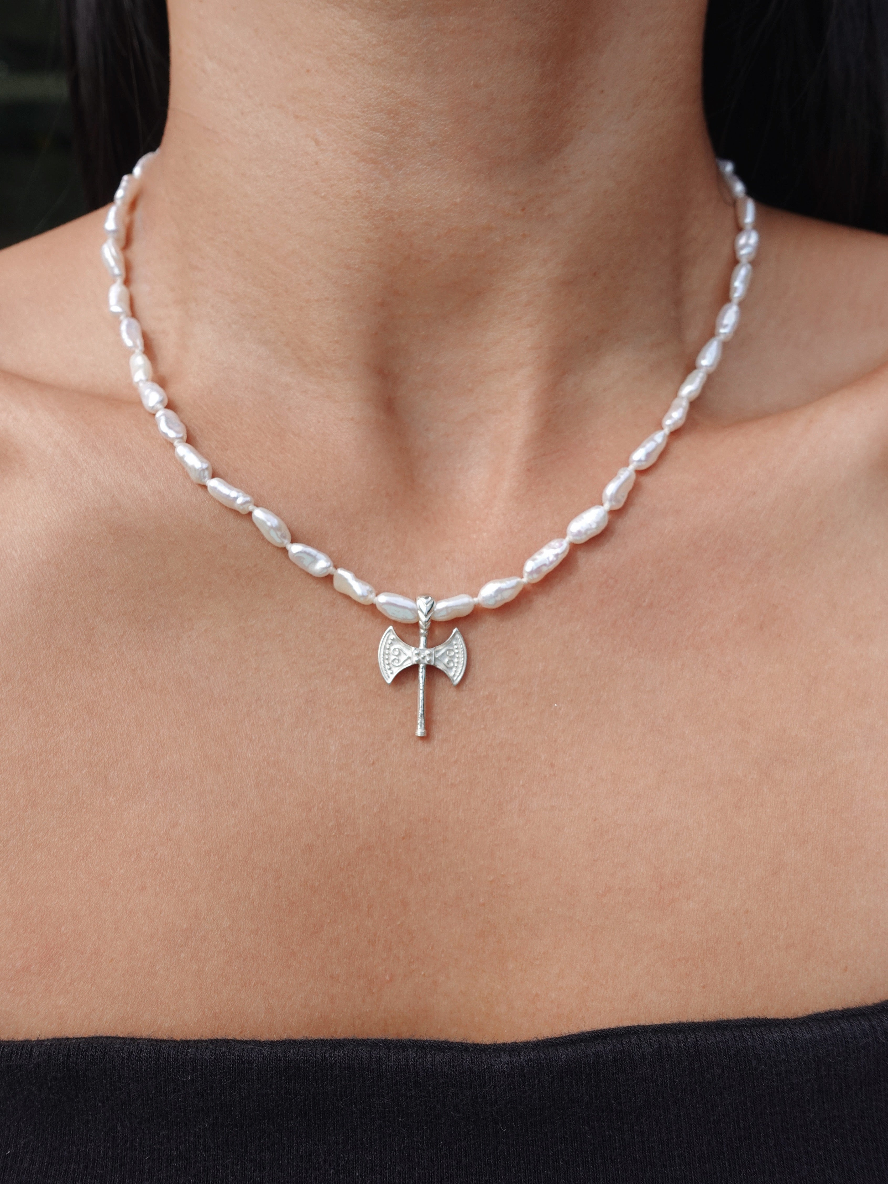 A close-up of a person's neck and upper chest wearing a silver necklace with irregular white pearl-like beads and a small axe pendant at the center.