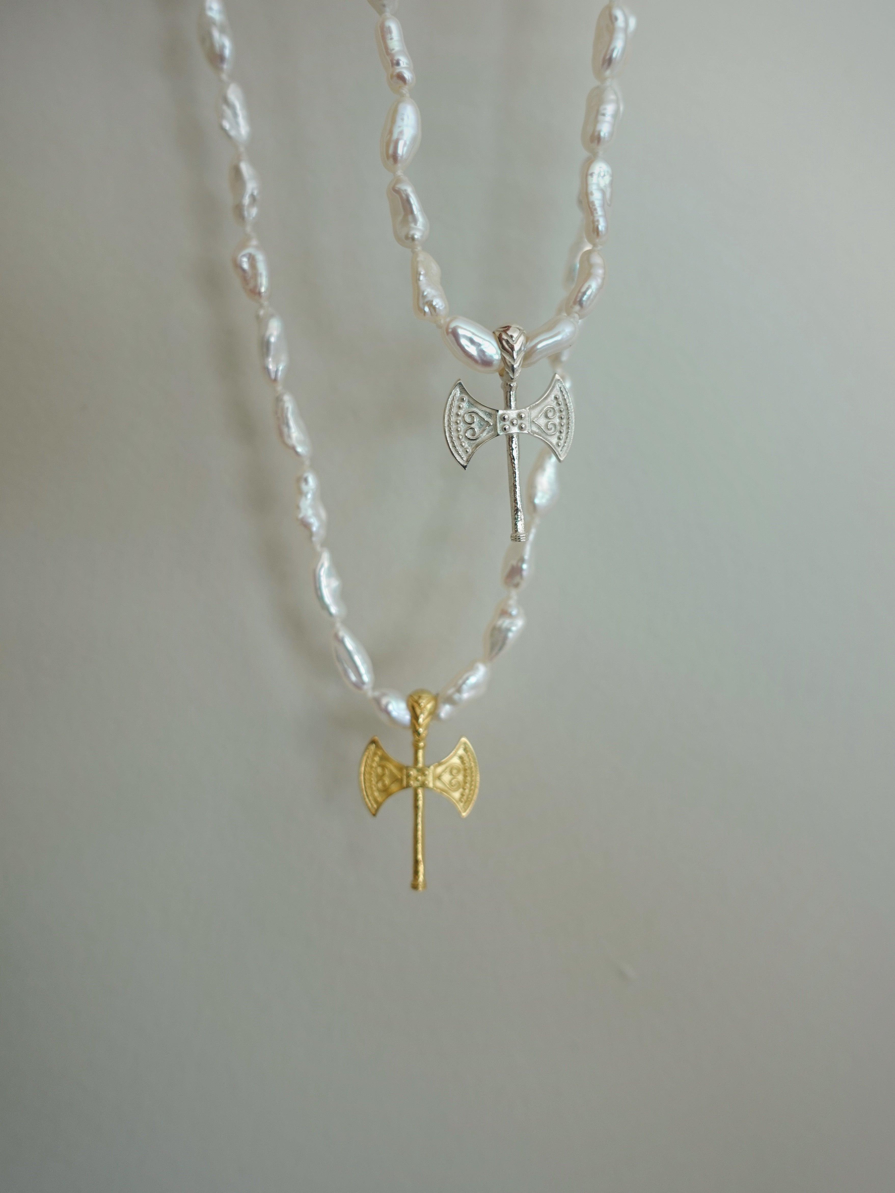 A double-strand necklace featuring white shell beads and two axe pendants, one in textured gold and one in silver with small clear stones, displayed against a plain light gray background.