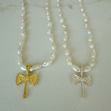 Two beaded necklaces with small axe pendants displayed on a light marble surface. One necklace has a gold axe pendant, and the other has a silver axe pendant.