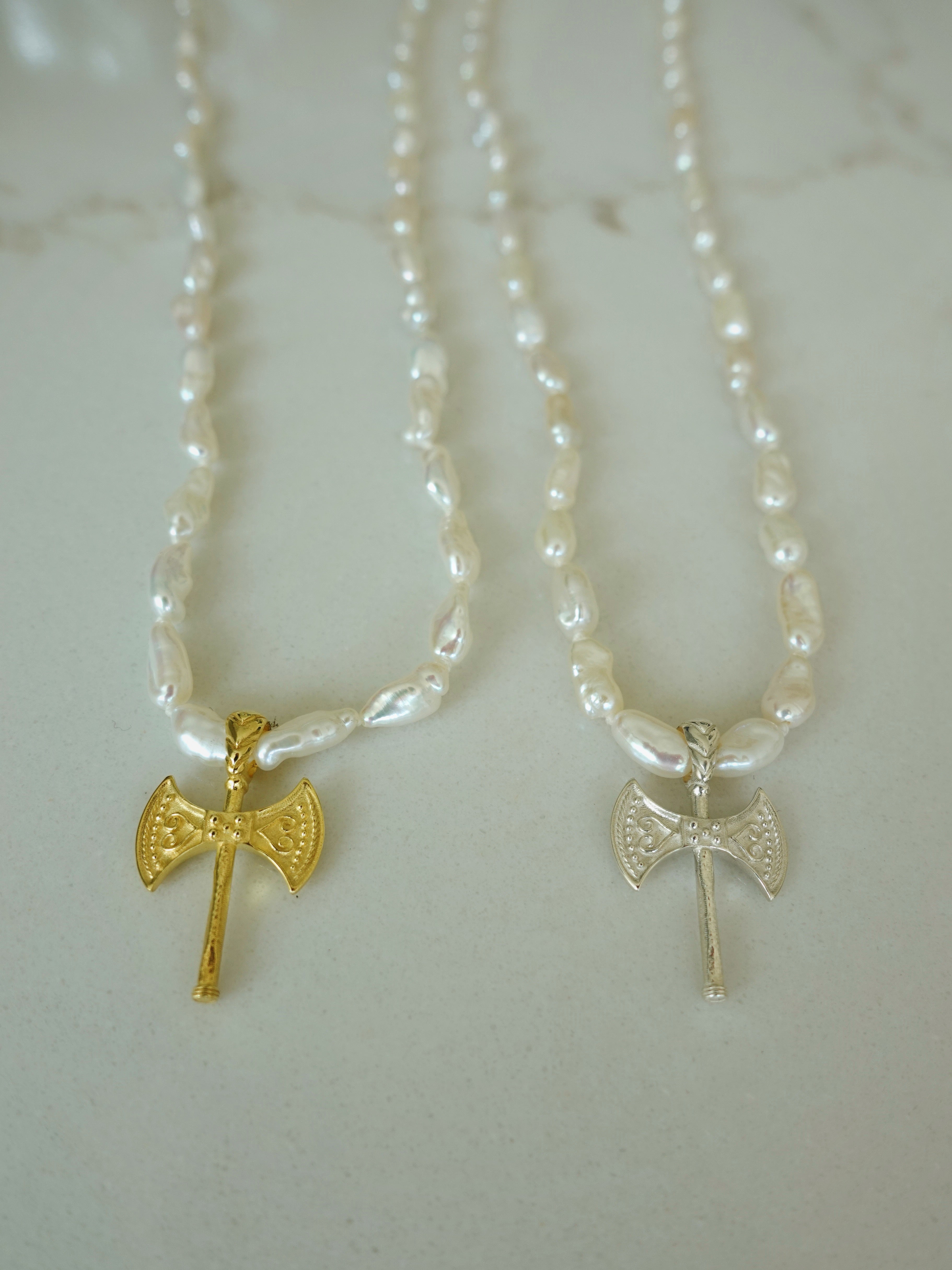 Two beaded necklaces with small axe pendants displayed on a light marble surface. One necklace has a gold axe pendant, and the other has a silver axe pendant.