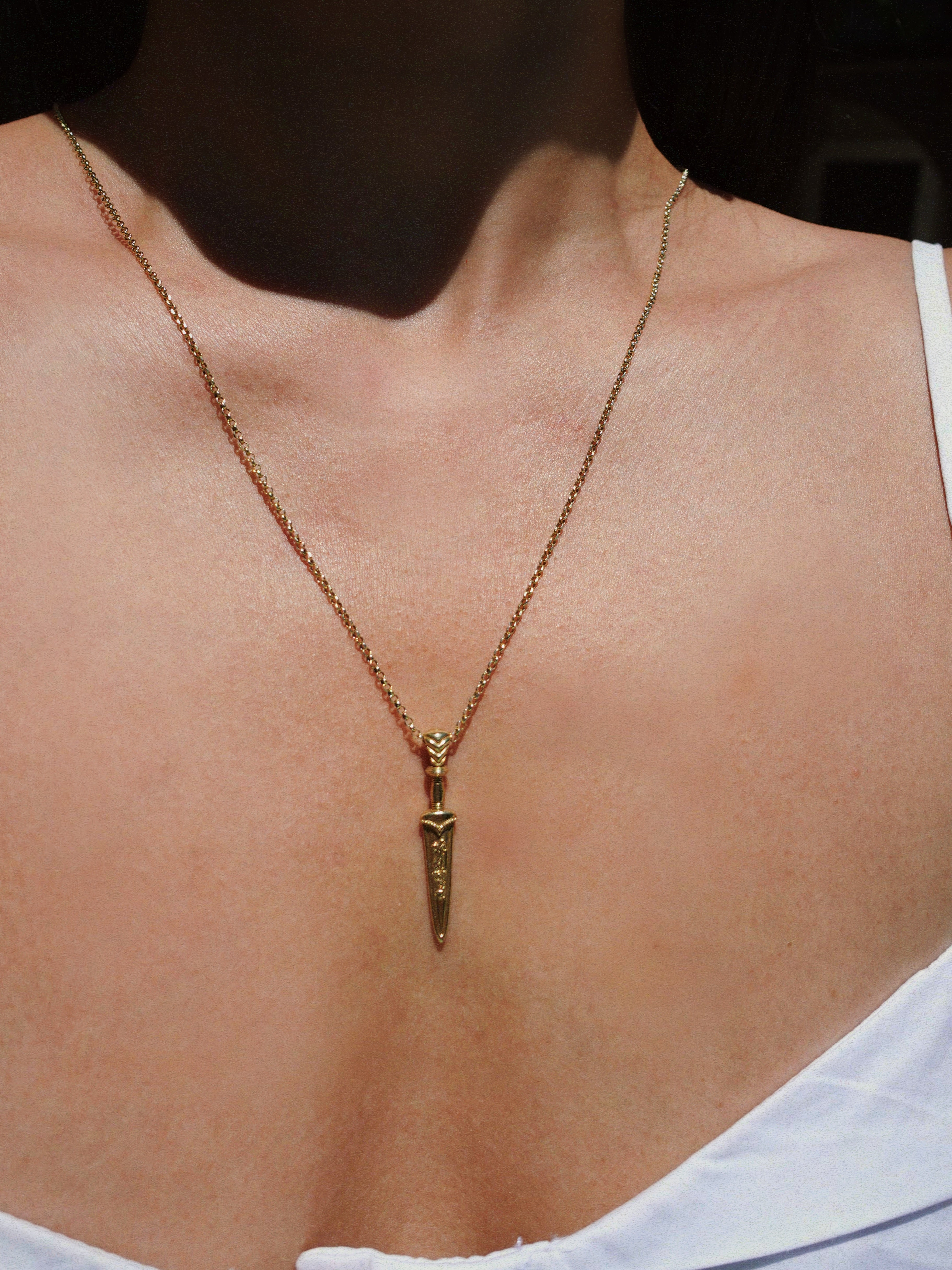 Gold necklace with a sword-shaped pendant on a person wearing a white top.