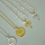 Four delicate necklaces arranged diagonally on a light gray surface, featuring white baroque pearls, yellow gold, and silver metal chains with round medallion pendants and small angel wing charms.