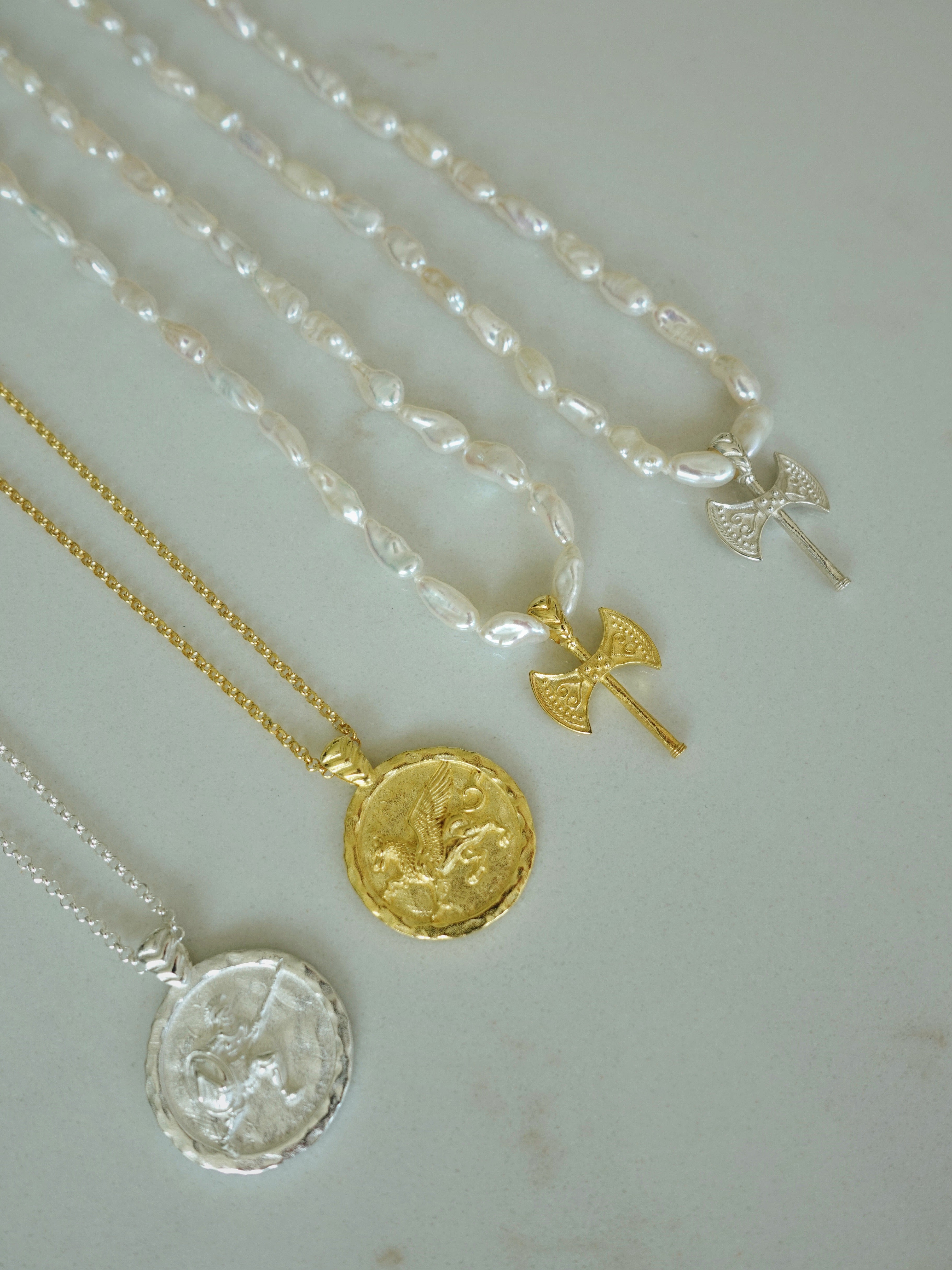 Four delicate necklaces arranged diagonally on a light gray surface, featuring white baroque pearls, yellow gold, and silver metal chains with round medallion pendants and small angel wing charms.