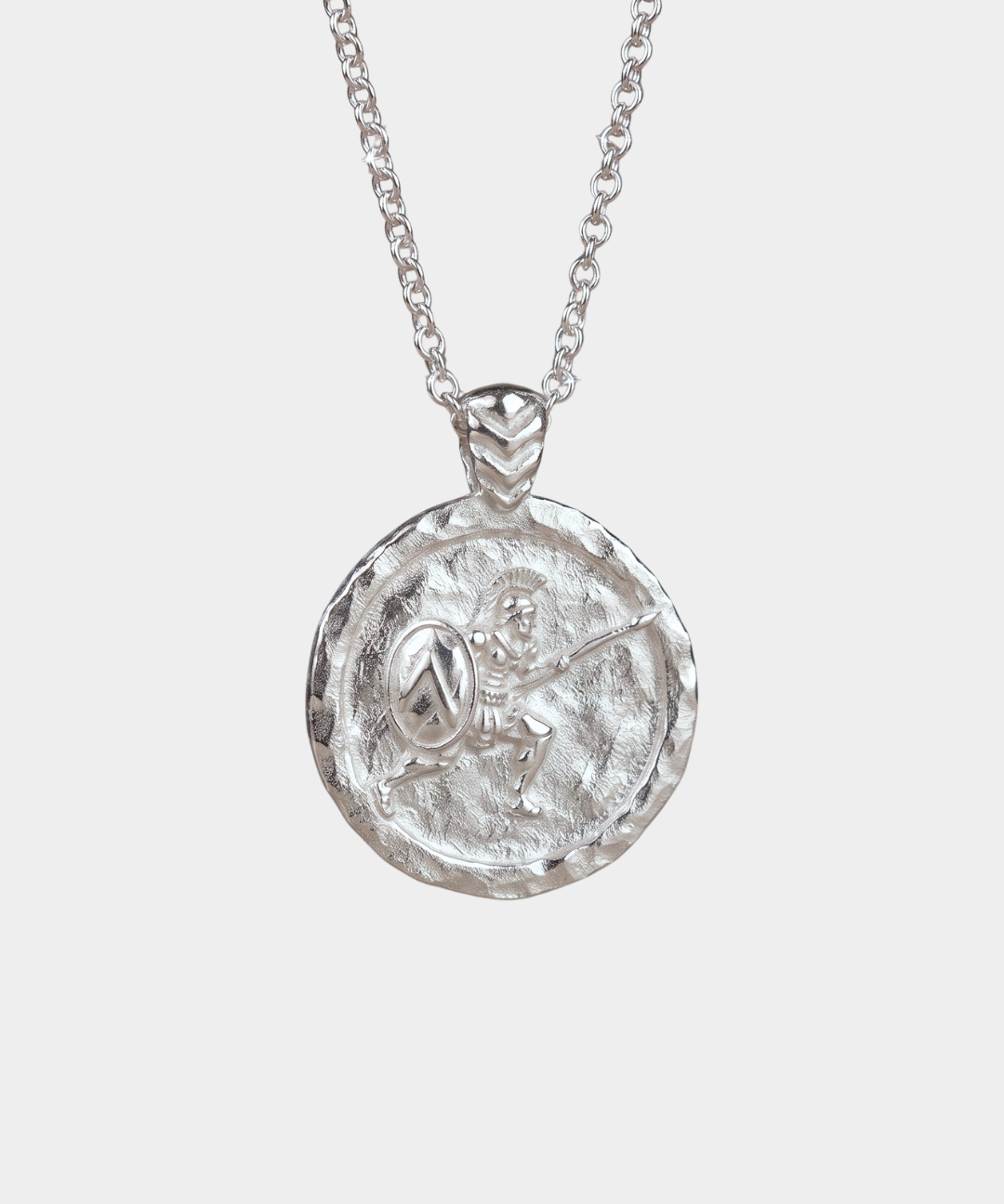 A silver pendant necklace featuring a circular, hammered-texture coin-style medallion with an embossed figure like ancient greek warrior holding a shield, suspended from a delicate silver chain against a light gray background.