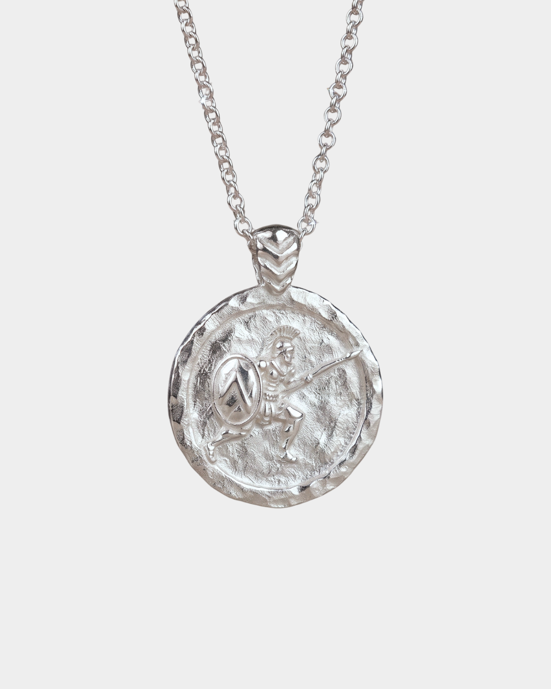 A silver pendant necklace featuring a circular, hammered-texture coin-style medallion with an embossed figure like ancient greek warrior holding a shield, suspended from a delicate silver chain against a light gray background.