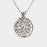 A silver pendant necklace featuring a hammered circular medallion with a textured organic surface and a small central star-like detail, suspended from a delicate silver chain against a light gray background.